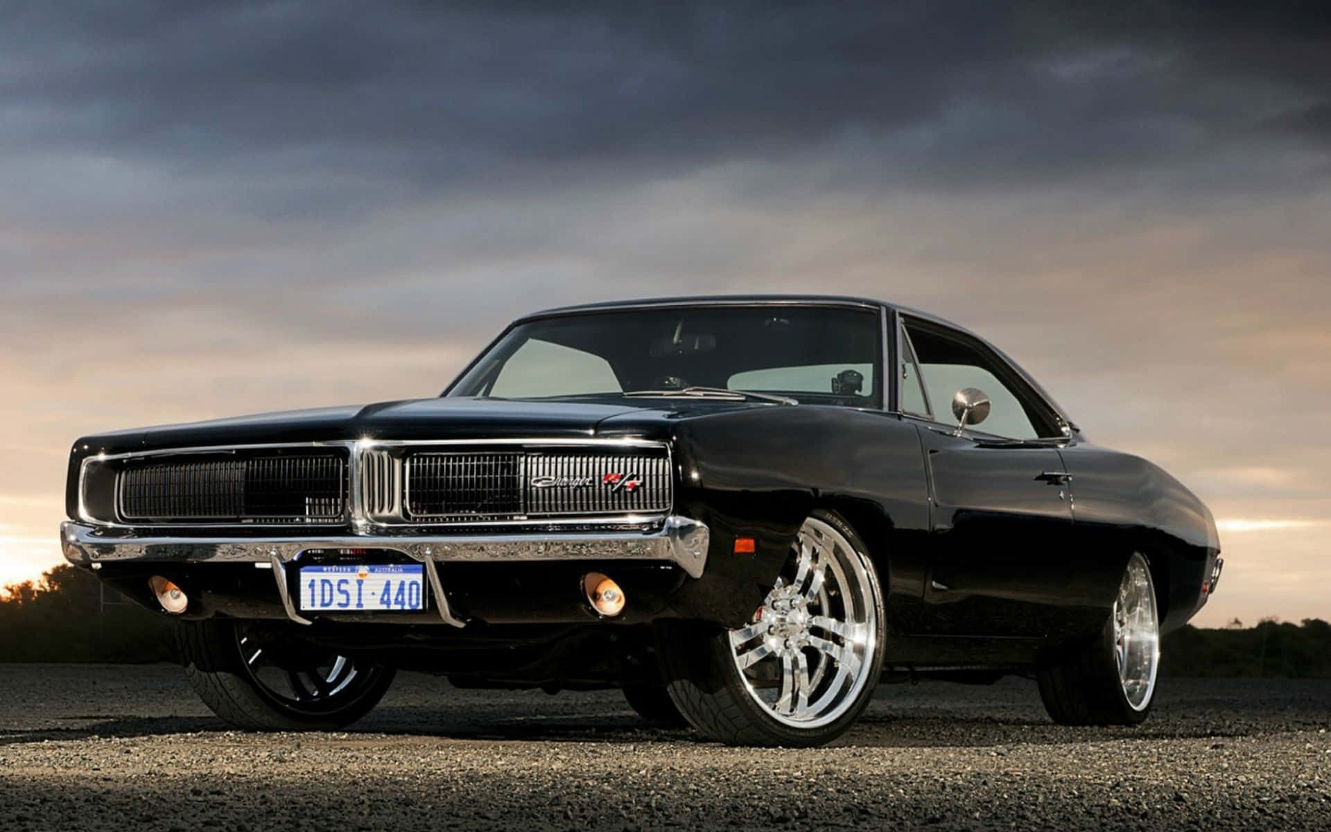 Dodge Charger 桌布 壁紙 — 一輛光澤黑色經典肌肉車，帶有鍍鉻裝飾和定制輪圈，停在碎石路上，背景是戲劇性的暮色天空。