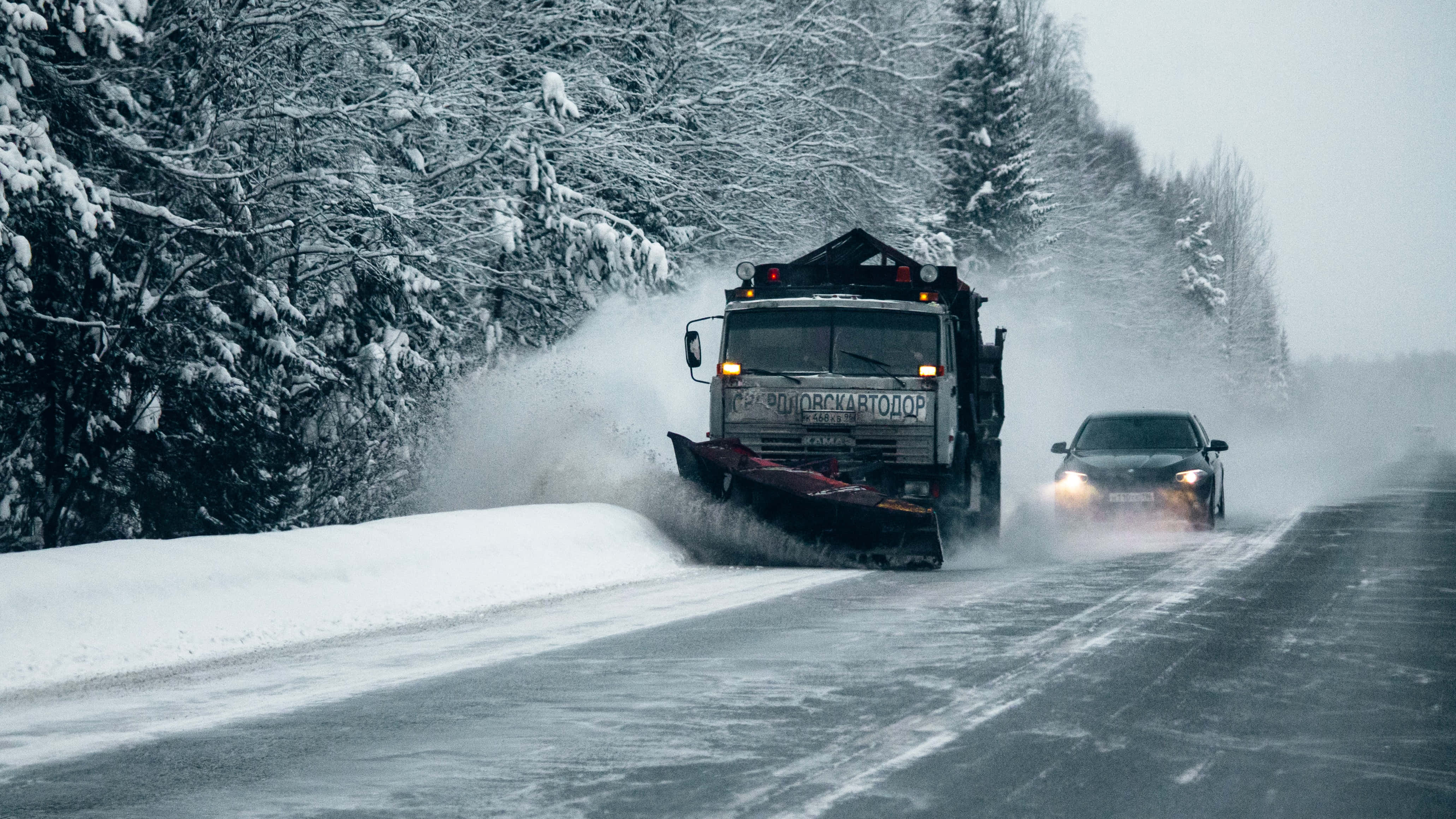 Snowplow 桌布 壁紙 — 一輛剷雪車正在清理一條被雪覆蓋的道路，道路兩旁是積雪的樹木，一輛汽車在冬季風暴中跟在後面。