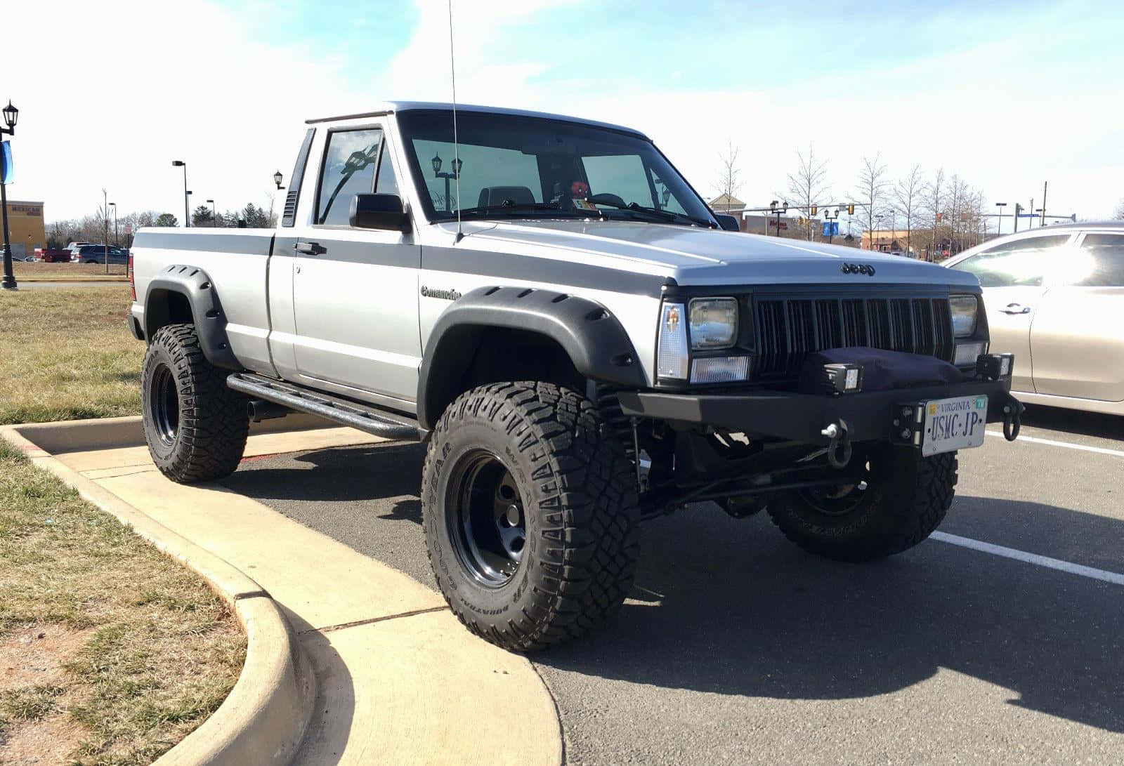 Jeep Comanche 桌布 壁紙 — 一輛升高了的銀色Jeep Cherokee皮卡車，配備超大輪胎，在陽光明媚的日子停泊在鋪砌的停車場。