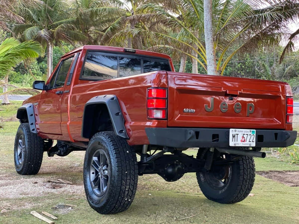 Jeep Comanche 桌布 壁紙 — 一輛升高嘅紅色Jeep Wrangler，配有大輪胎，停泊喺熱帶環境中嘅棕櫚樹下草地上。