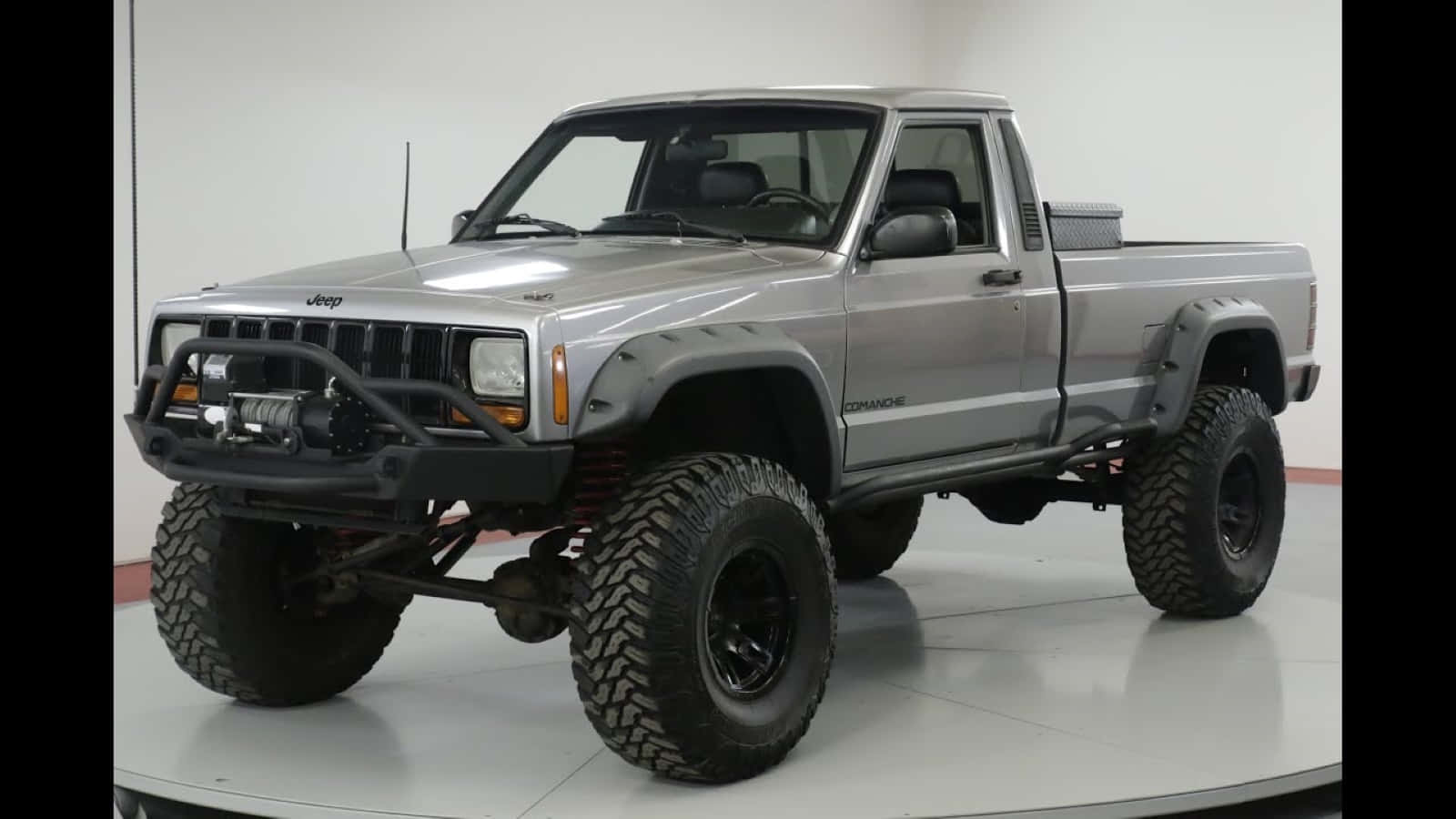 Jeep Comanche 桌布 壁紙 — 一輛升高嘅銀色Jeep Cherokee XJ，配備超大輪胎同黑色保險槓，企喺白色嘅工作室轉盤上。