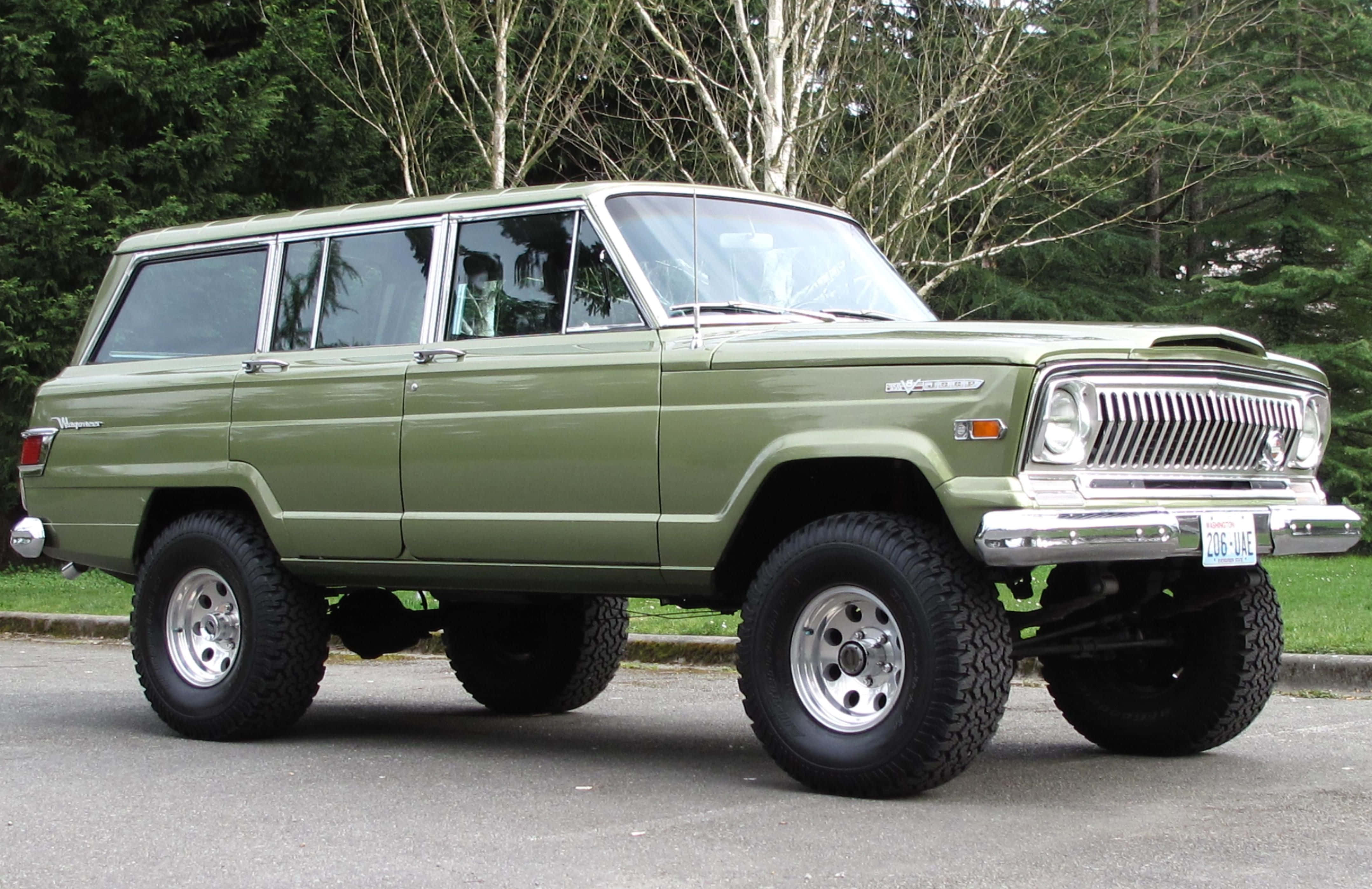 Jeep Wagoneer 桌布 壁紙 — 一輛升高底盤的橄欖綠色經典Jeep Wagoneer，配備鍍鉻格柵和大輪胎，停在樹旁的瀝青路面上。