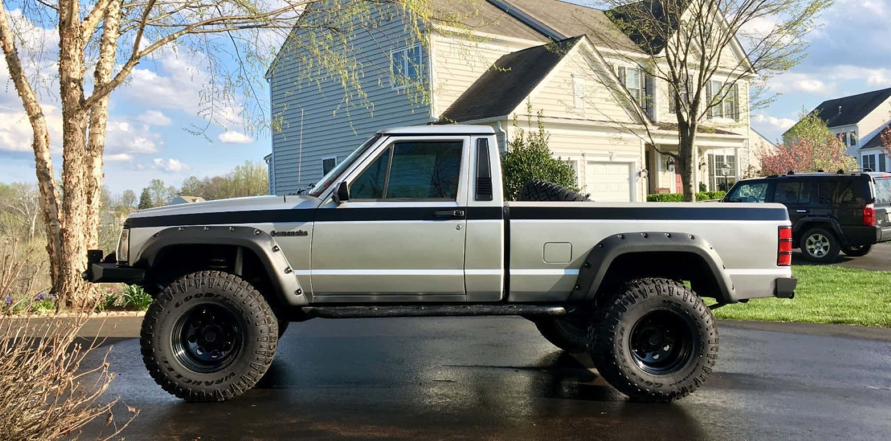 Jeep Comanche 桌布 壁紙 — 一輛升高底盤的銀色Jeep Cherokee皮卡車，配備超大輪胎，停放在郊區房屋前的濕車道上。