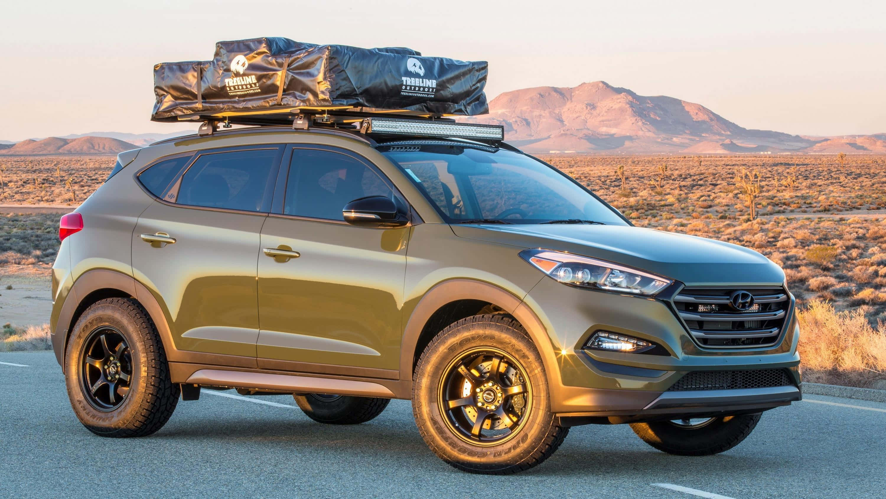 Hyundai Tucson 桌布 壁紙 — 一輛啞光橄欖綠色的SUV，車頂裝有行李袋和黑色輪圈，在黃金時段停泊在沙漠公路上。