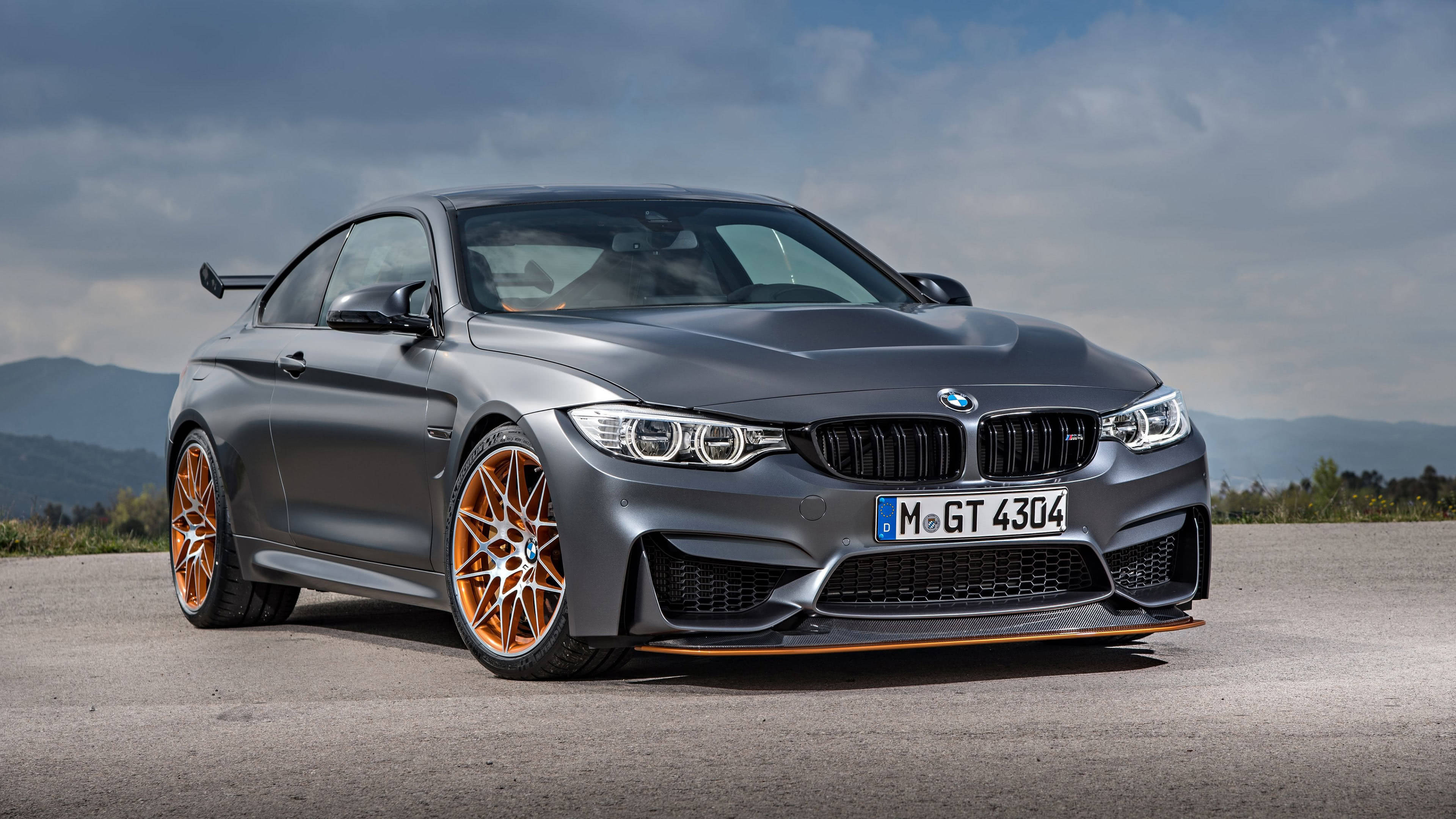 4K BMW M4 GTS 壁紙 — 一輛啞光灰色BMW M4 GT4跑車，配橙色輪圈，停泊在瀝青路上，背景是陰天和遠山。