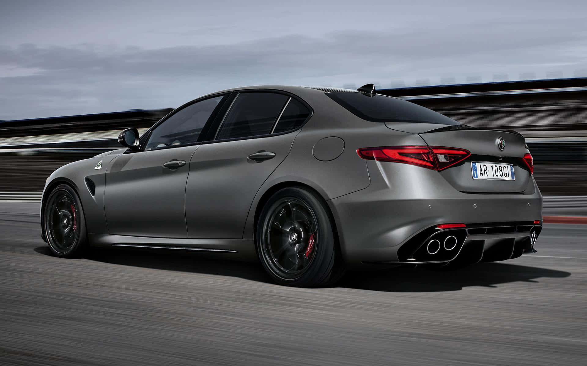 Alfa Romeo Giulia 桌布 壁紙 — 一輛啞光灰色的Alfa Romeo房車在多雲的天空下，在模糊的賽道上疾馳。
