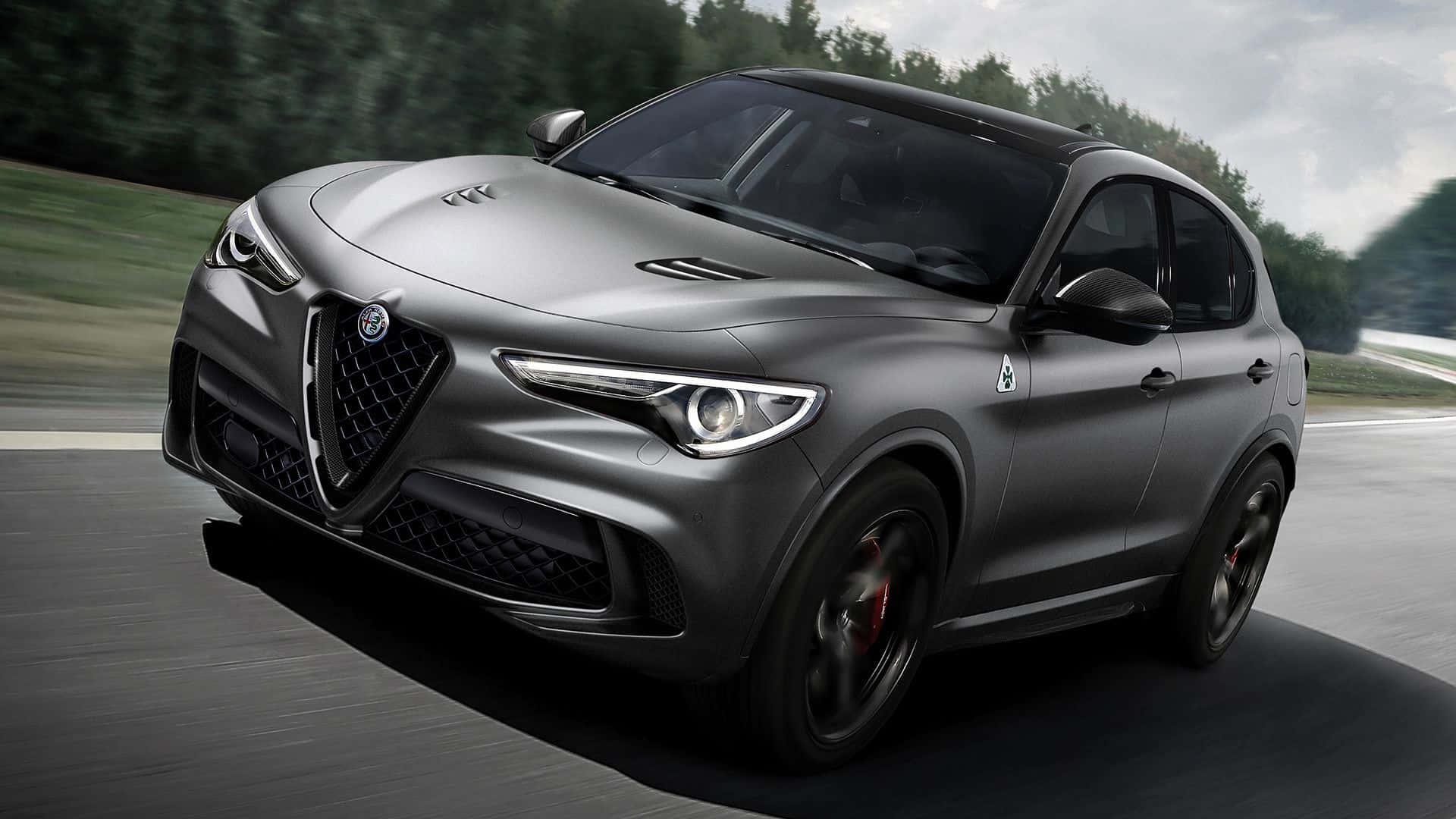 Alfa Romeo Stelvio 桌布 壁紙 — 一輛啞光灰色的Alfa Romeo Stelvio Quadrifoglio在蜿蜒的道路上疾馳，背景是模糊的綠色樹木。
