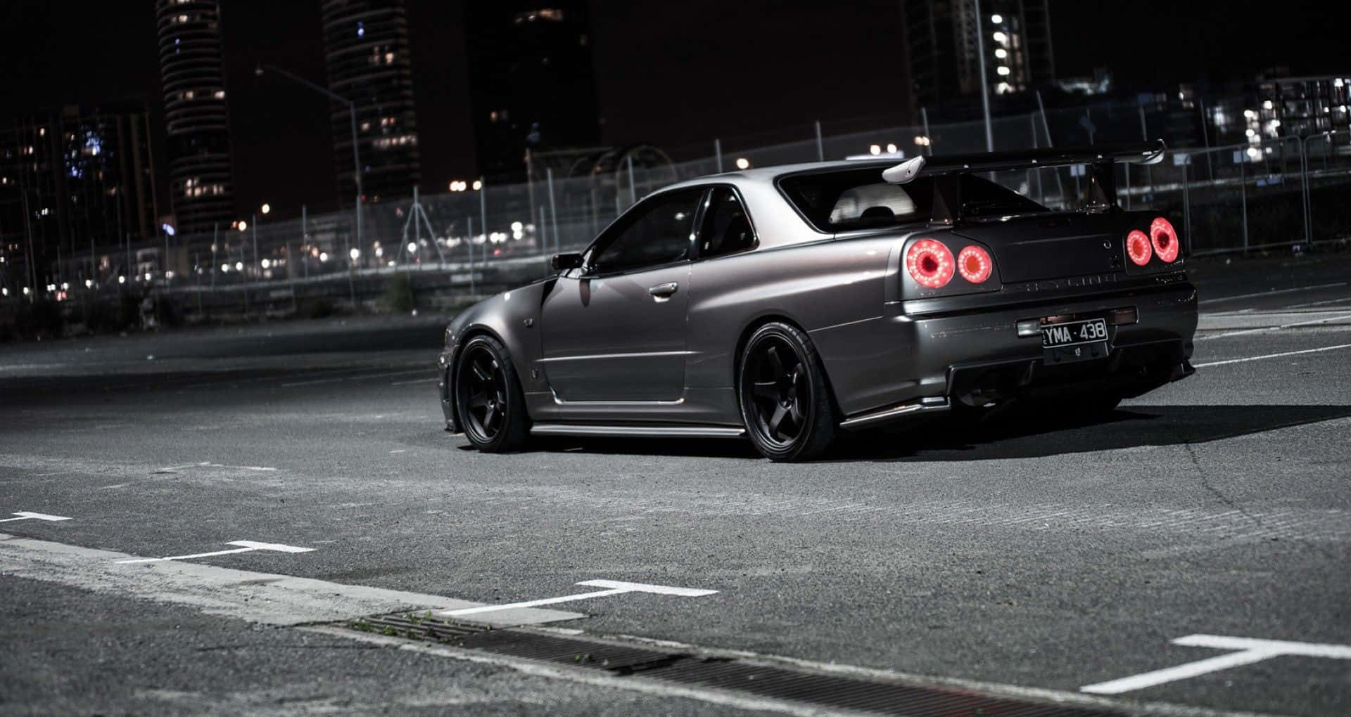 JDM 車 桌布 壁紙 — 一輛啞光灰色的日產Skyline R34跑車，帶有發光的紅色尾燈，夜晚停在空無一人的城市街道上。