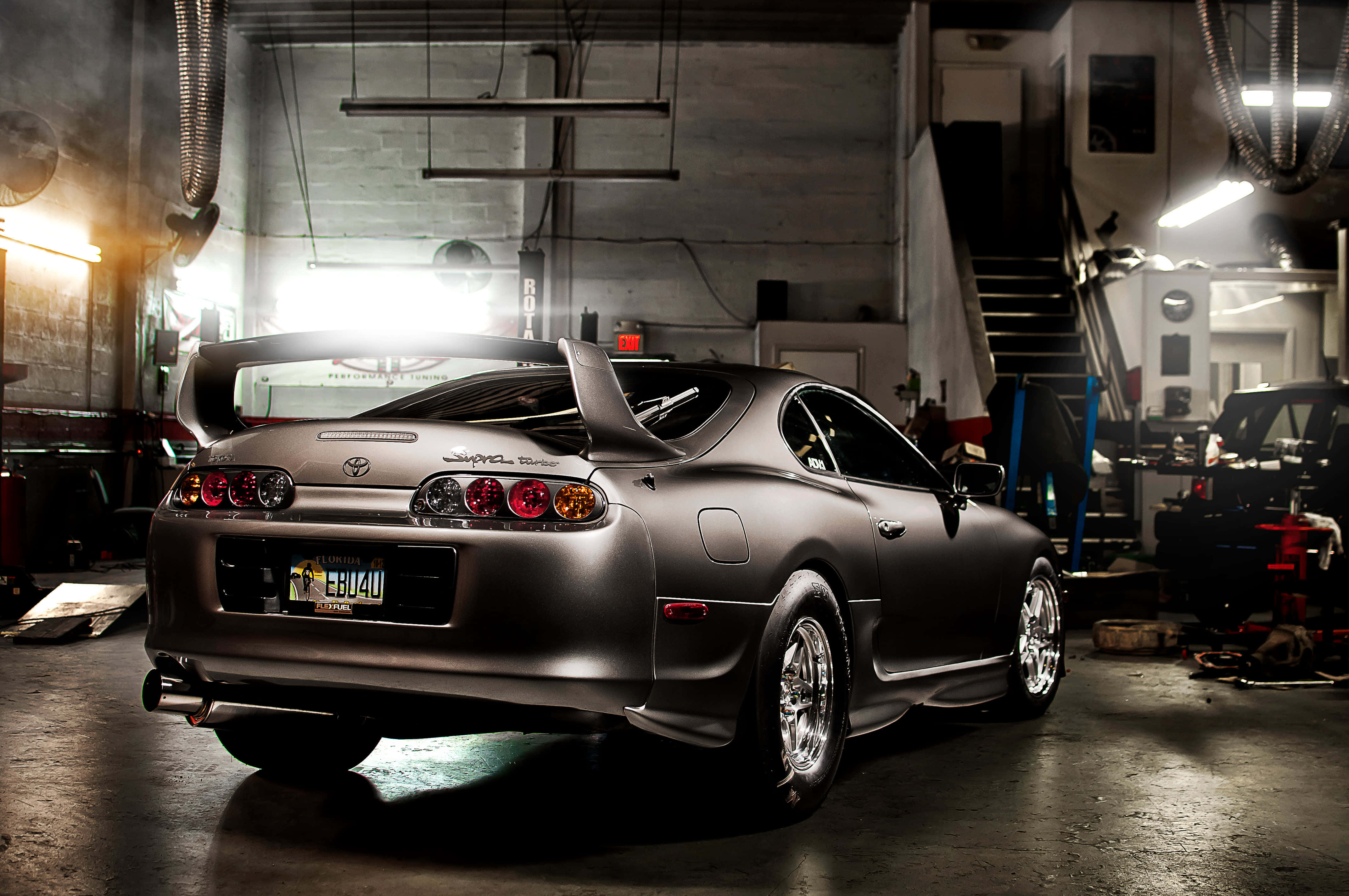 Supra 桌布 壁紙 — 一輛啞光灰色的豐田Supra跑車在車庫燈光下閃閃發光，煙霧在其後擾流板周圍飄散。