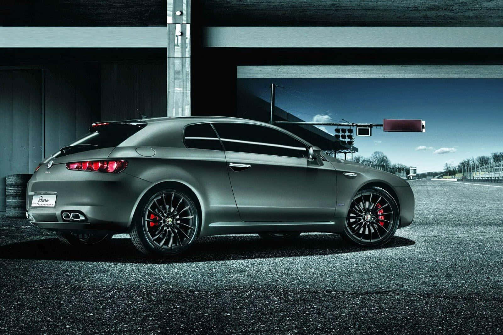 Alfa Romeo Brera 桌布 壁紙 — 一輛啞光灰色的阿爾法羅密歐跑車，配備黑色輪圈和紅色制動卡鉗，停泊在現代結構下的賽道上。