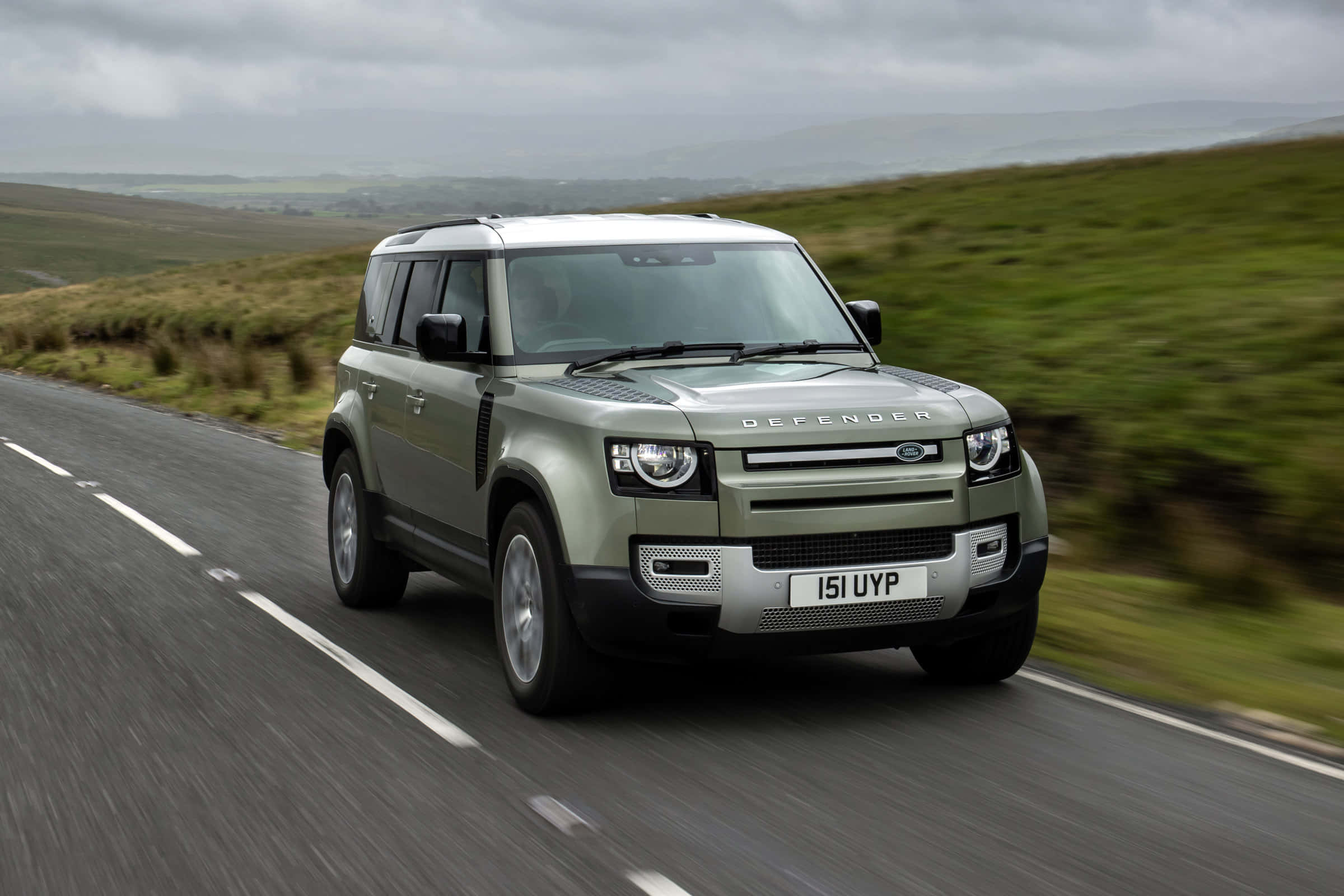 Land Rover Defender 桌布 壁紙 — 一輛啞光綠色的 Land Rover Defender 在陰天下一條蜿蜒的鄉村道路上疾馳，背景是模糊的綠色山丘。