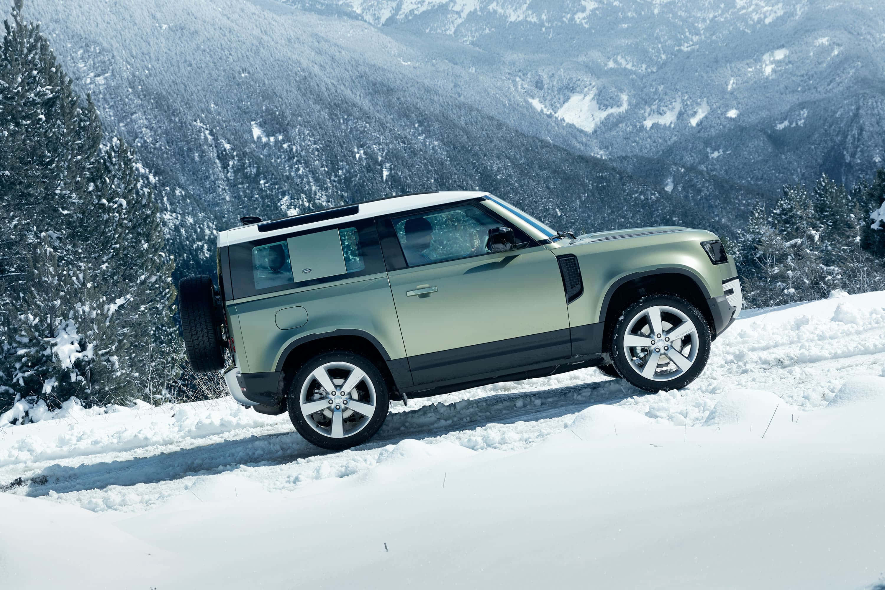 Land Rover Defender 桌布 壁紙 — 一輛啞光綠色的Land Rover Defender在積雪深厚的山路上行駛，兩旁是白雪覆蓋的松樹。