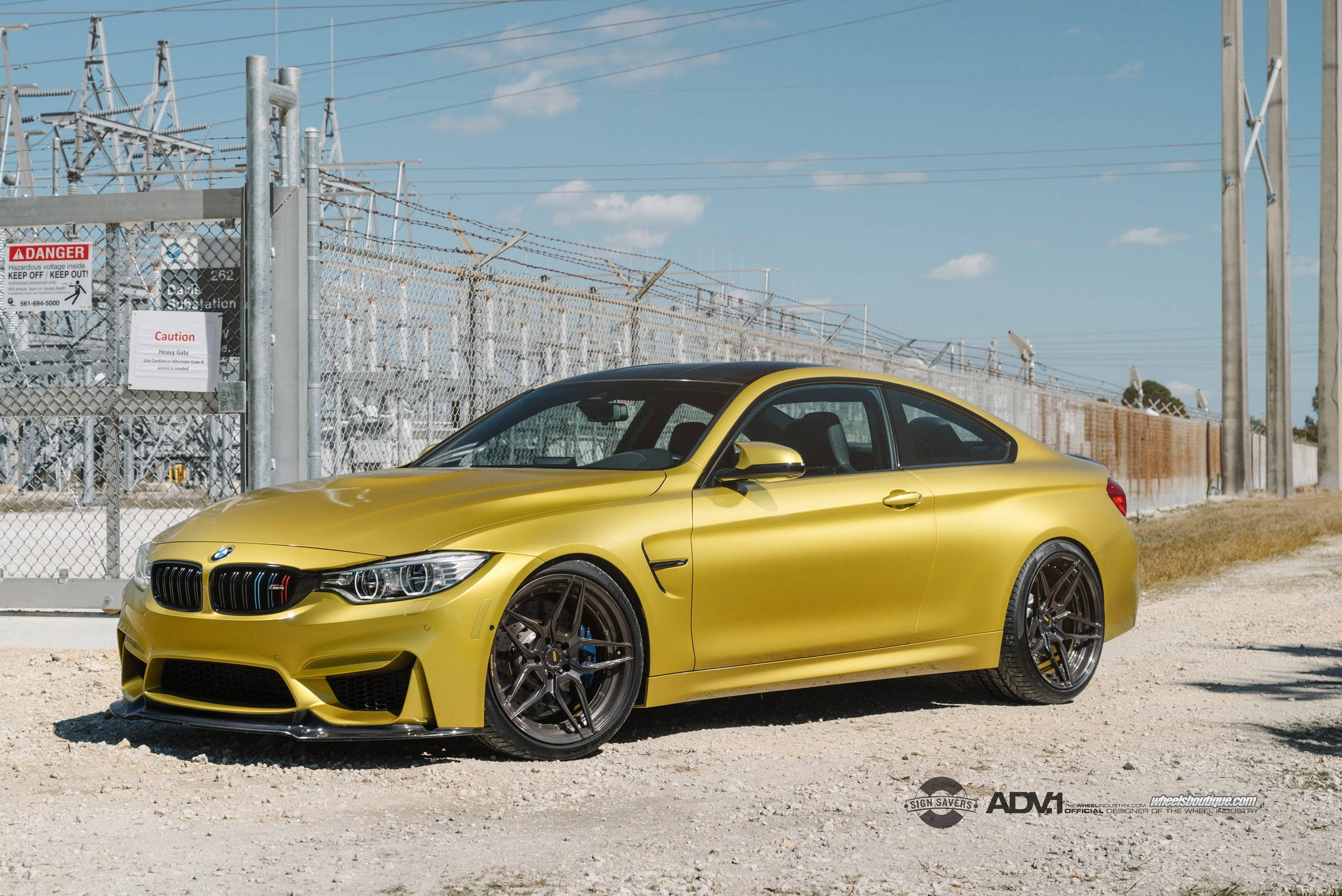 Sports Car 桌布 壁紙 — 一輛啞光金色的BMW M4雙門跑車停泊在碎石路上，旁邊是圍欄圍著的電力變電站，天空清澈湛藍。