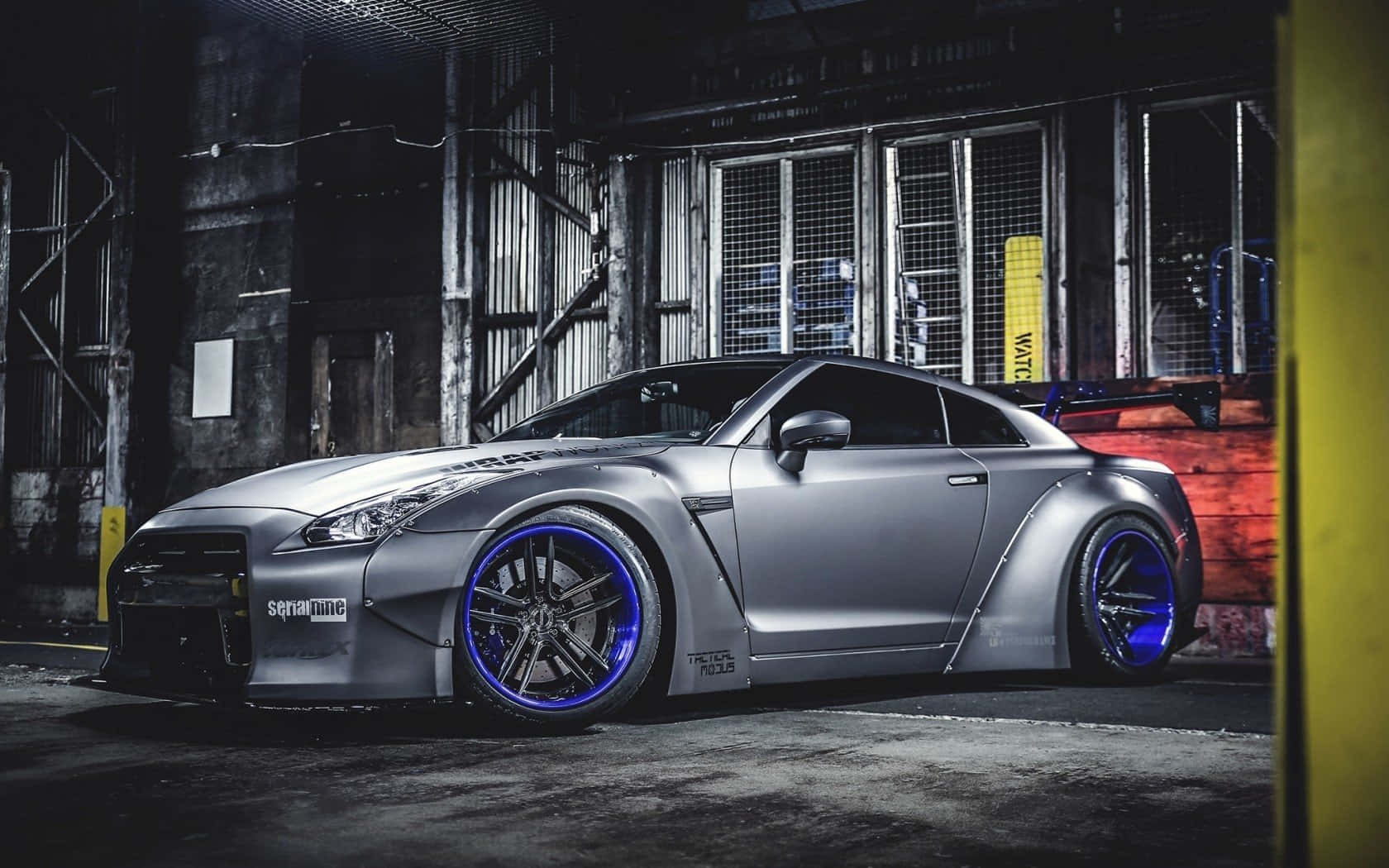 Gtr 桌布 壁紙 — 一輛啞光銀色日產GT-R配備發光藍色輪圈，夜晚停泊在粗糙的工業倉庫中。