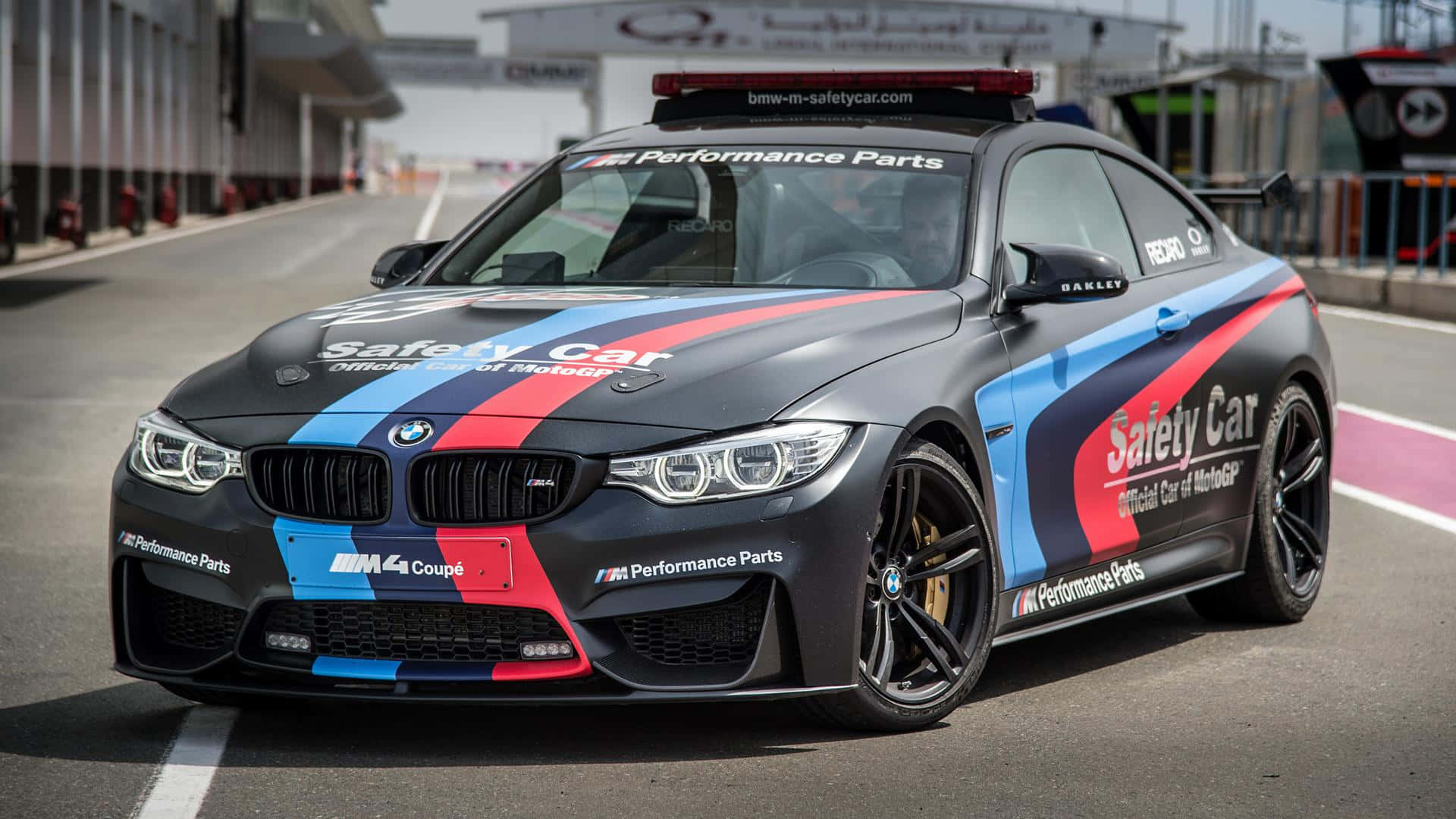 Safety Car 桌布 壁紙 — 一輛啞光黑色BMW M4 Coupe安全車，帶有藍、紅、白色賽車條紋，停在賽道上。