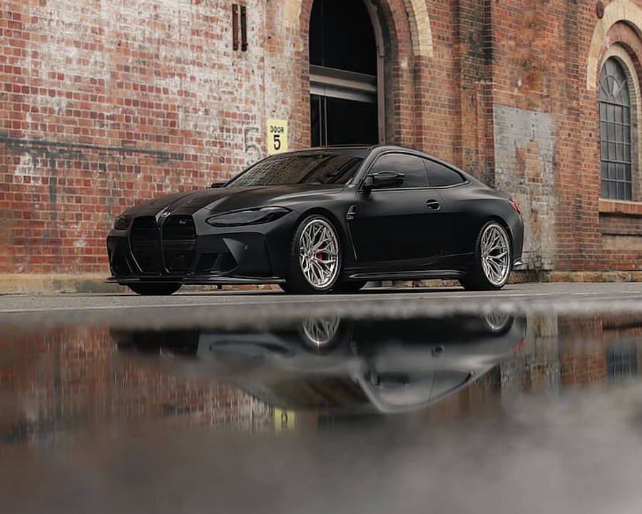 Bmw M4 桌布 壁紙 — 一輛啞光黑色BMW M4轎跑車在潮濕的路面上反射，旁邊是帶有拱形窗戶的風化磚倉庫。