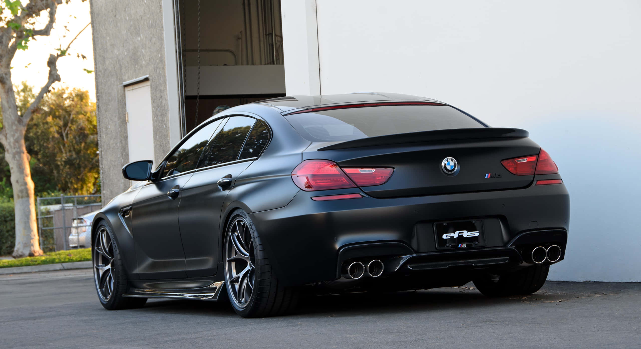Bmw M6 桌布 壁紙 — 一輛啞光黑色BMW M6 Gran Coupe，配備四出排氣管和紅色尾燈，在黃金時段停泊在混凝土建築物旁。