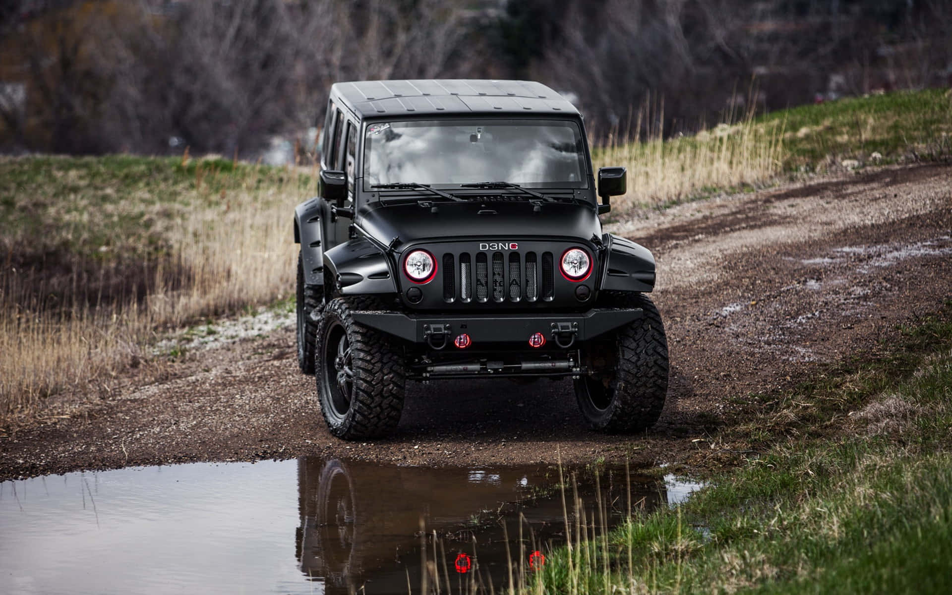 Jeep 動圖 桌布 壁紙 — 一輛啞光黑色Jeep Wrangler帶紅色點綴，在泥濘水坑中行駛，周圍是乾草和光禿禿的樹木。