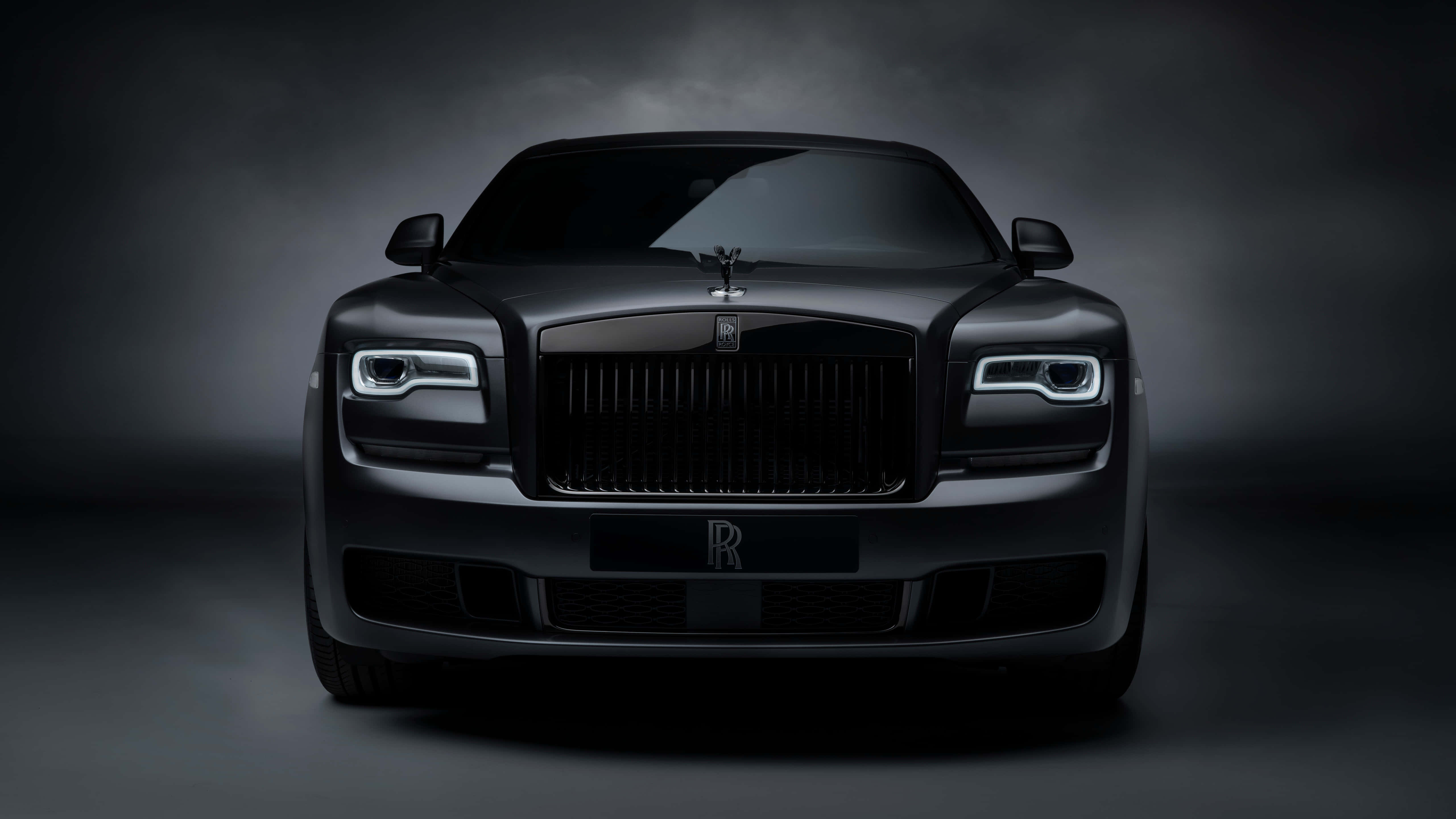 Rolls Royce 桌布 壁紙 — 一輛啞光黑色勞斯萊斯幻影的前視圖，在煙霧瀰漫的暗色工作室背景下，銳利的LED頭燈閃耀著光芒。