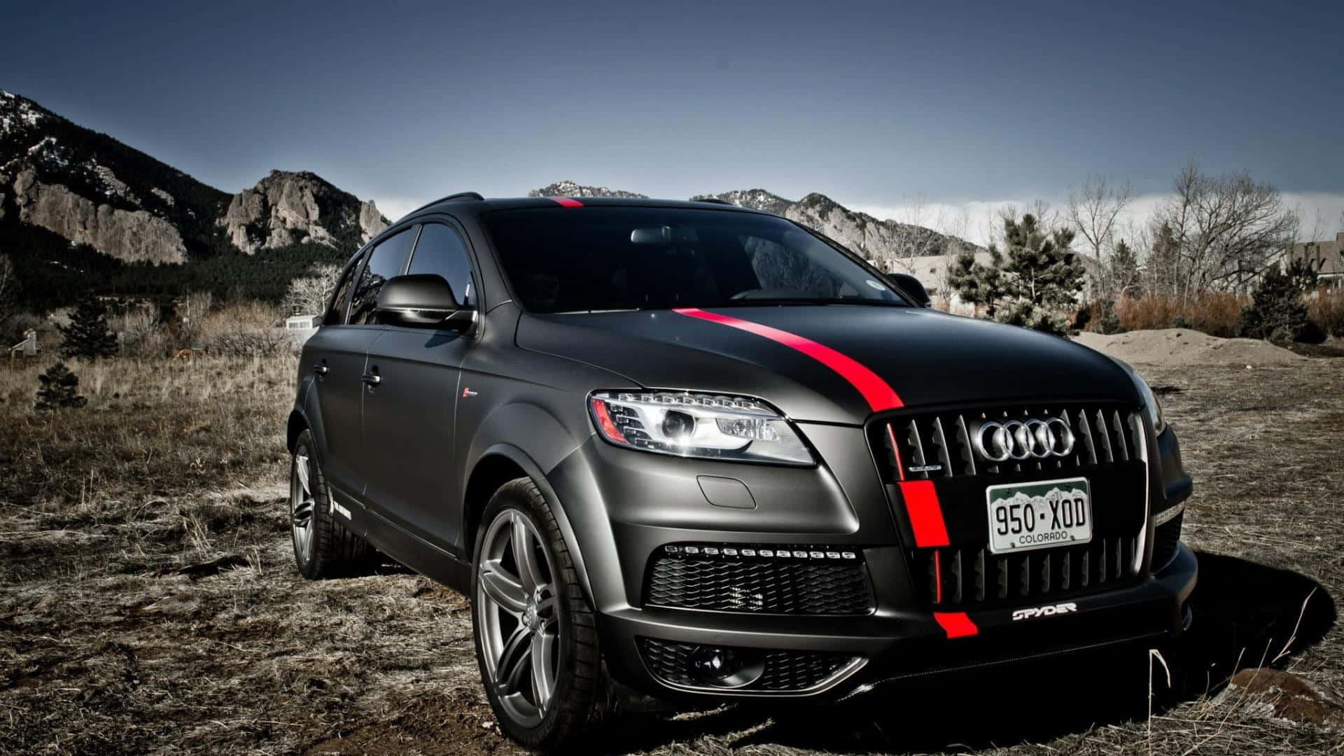 Audi Q7 桌布 壁紙 — 一輛啞光黑色奧迪Q7，帶有醒目的紅色賽車條紋，停在乾燥的草地上，背景是岩石山脈和清澈的藍天。