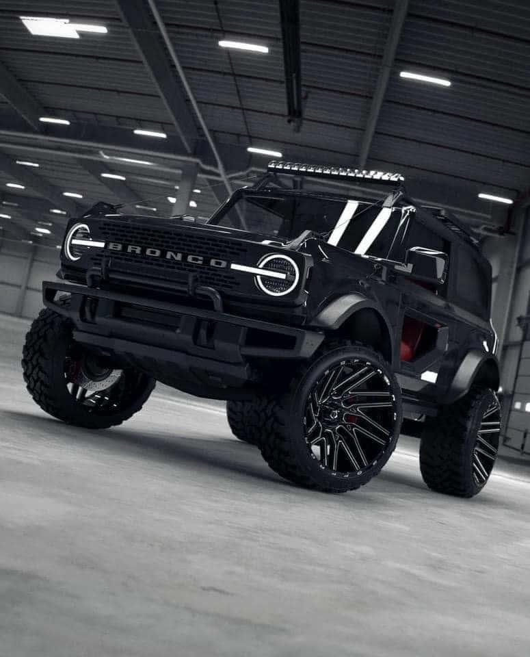 Off-road Car 桌布 壁紙 — 一輛啞光黑色定制Bronco SUV，配備超大輪圈和車頂燈，停放在寬敞的工業車庫中，上方有明亮的頂燈。