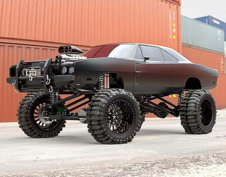 Off-road Car 桌布 壁紙 — 一輛啞光黑色怪獸卡車，配備超大輪胎和裸露懸掛系統，停在堆疊的貨運集裝箱前。
