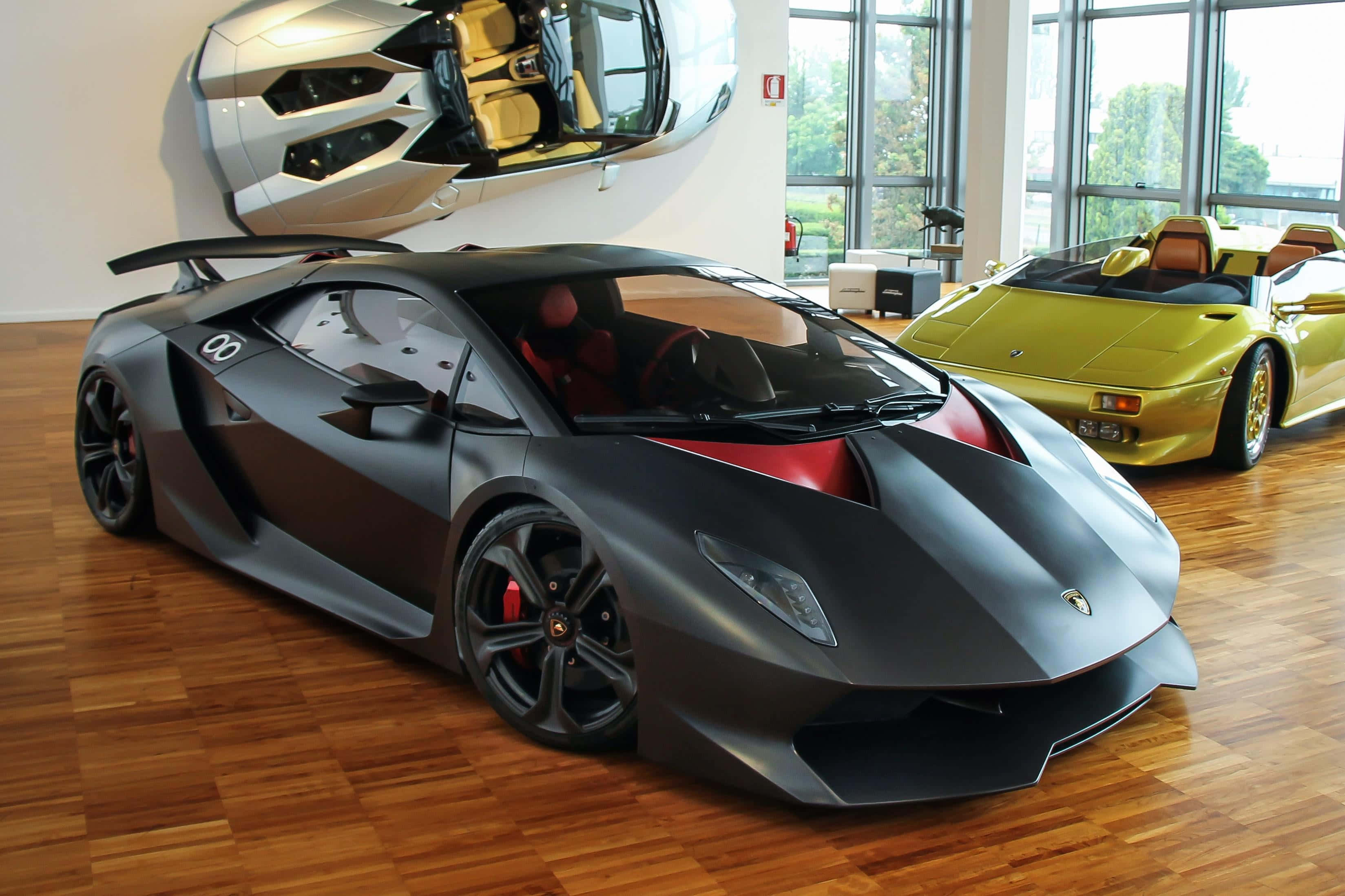 Lamborghini Sesto Elemento 桌布 壁紙 — 一輛啞光黑色林寶堅尼跑車帶有紅色點綴，突出地放置在明亮展廳的木地板上，背景有一輛黃色開篷車和一個壁掛式汽車雕塑。