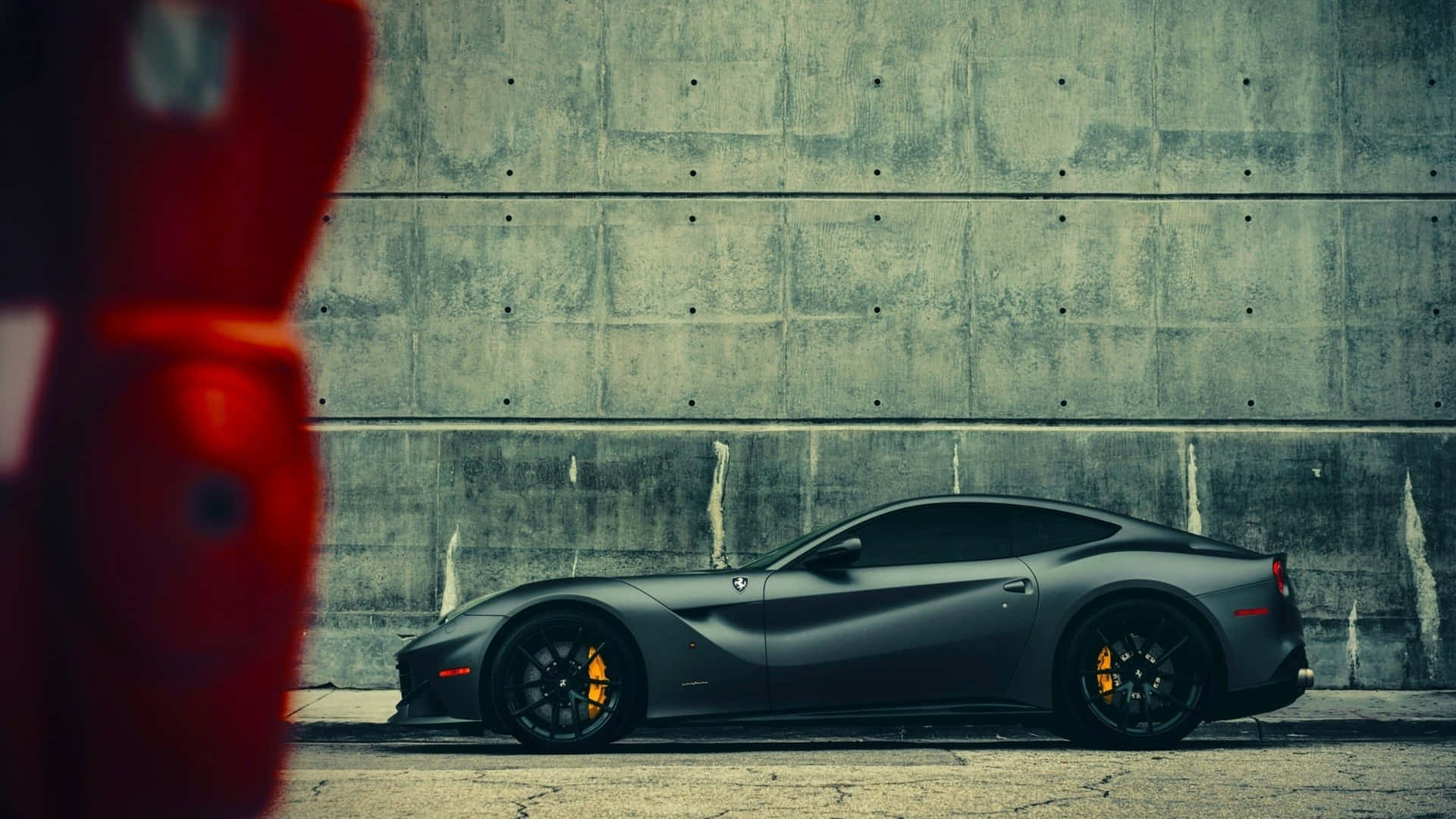 Ferrari F12 Berlinetta 桌布 壁紙 — 一輛啞光黑色法拉利跑車停泊在風化的混凝土牆前，前景有一個模糊的紅色物體。