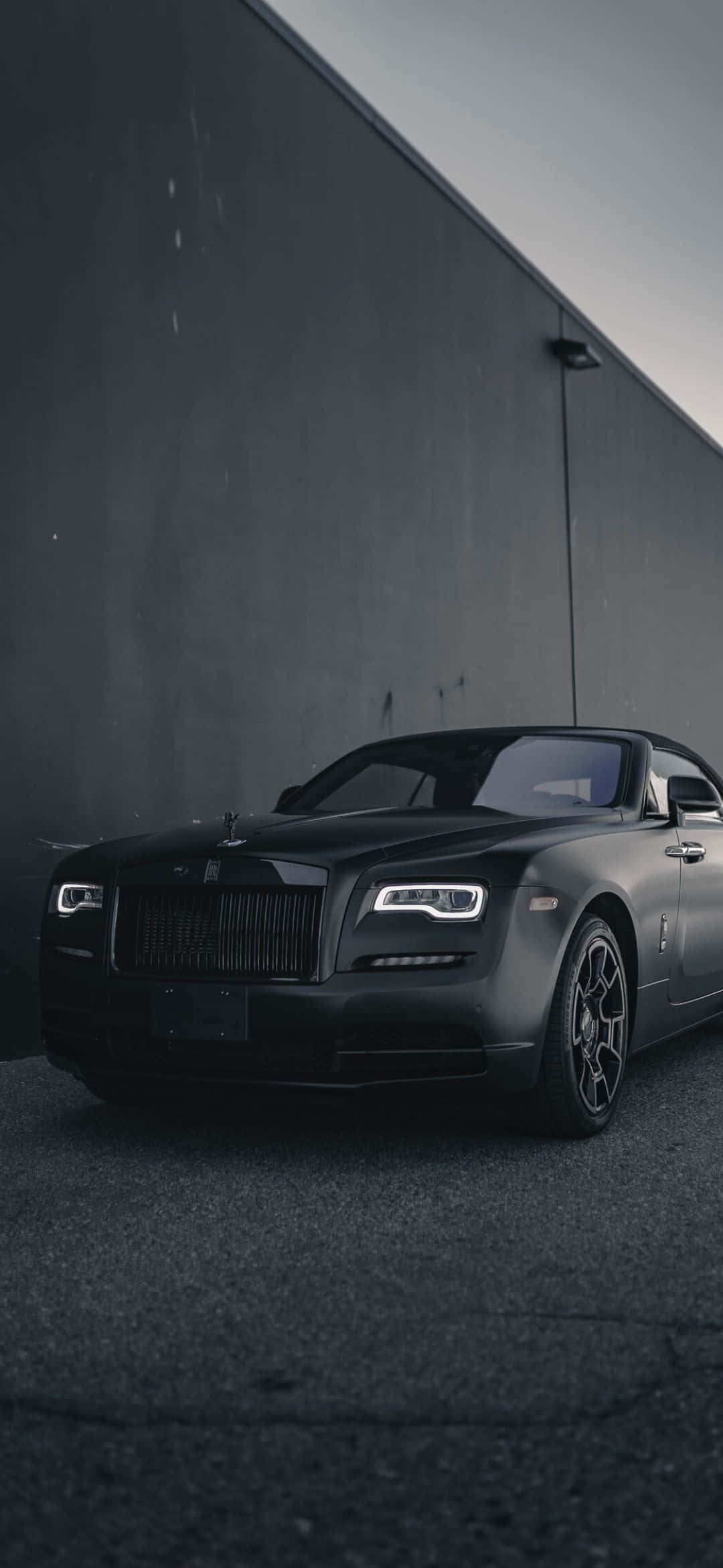 Rolls Royce 桌布 壁紙 — 一輛啞光黑色的勞斯萊斯在陰沉天空下，優雅地停靠在高聳的灰色混凝土牆旁。