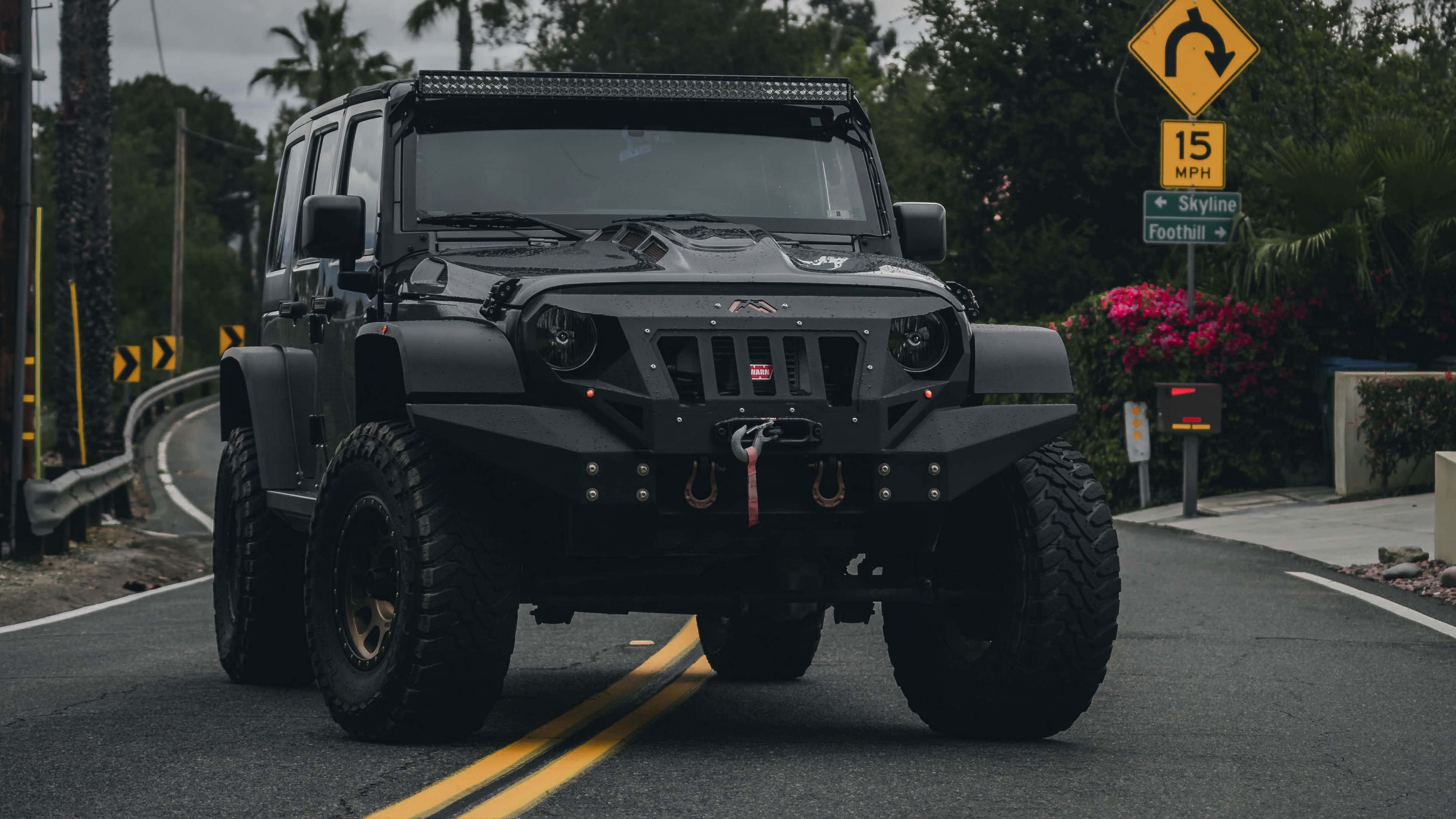 Jeep Cj 桌布 壁紙 — 一輛啞光黑色的越野Jeep Wrangler配備超大輪胎，行駛在棕櫚樹和色彩繽紛的花朵環繞的蜿蜒道路上。