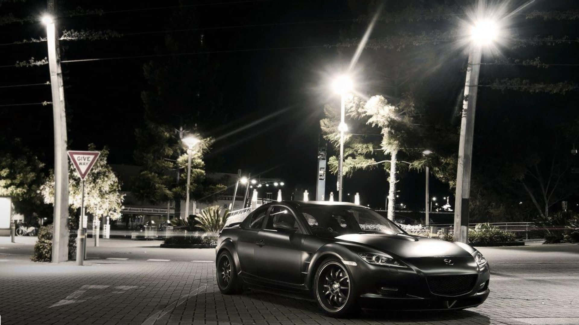 Mazda Rx-8 桌布 壁紙 — 一輛啞光黑色跑車在寧靜的夜間城市廣場上，在明亮的街燈下閃閃發光，周圍環繞著樹木和標誌。