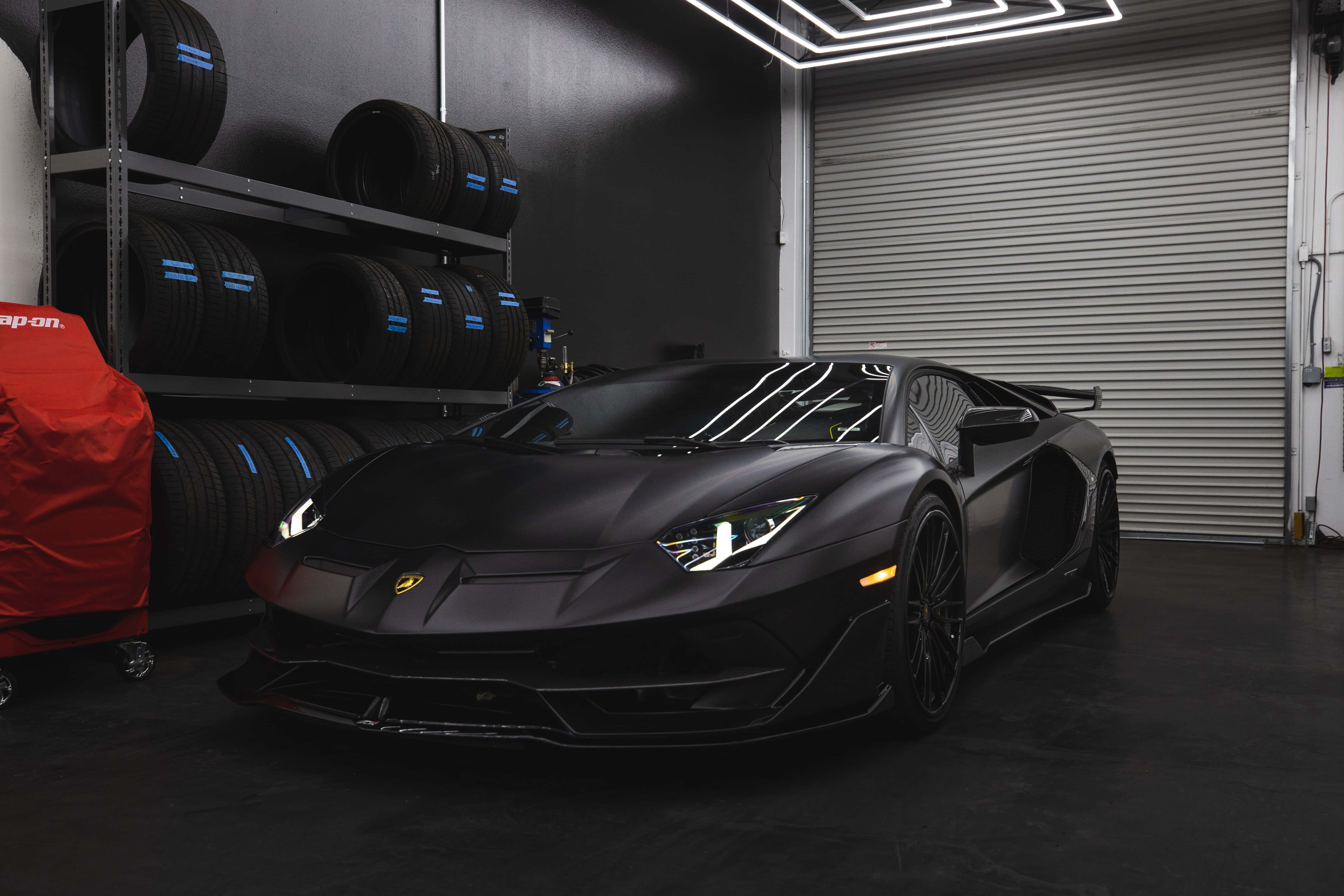 啞光黑色 桌布 壁紙 — 一輛啞黑色林寶堅尼 Aventador SVJ 停放在一個時尚現代、帶有堆疊輪胎和霓虹燈的車庫中。