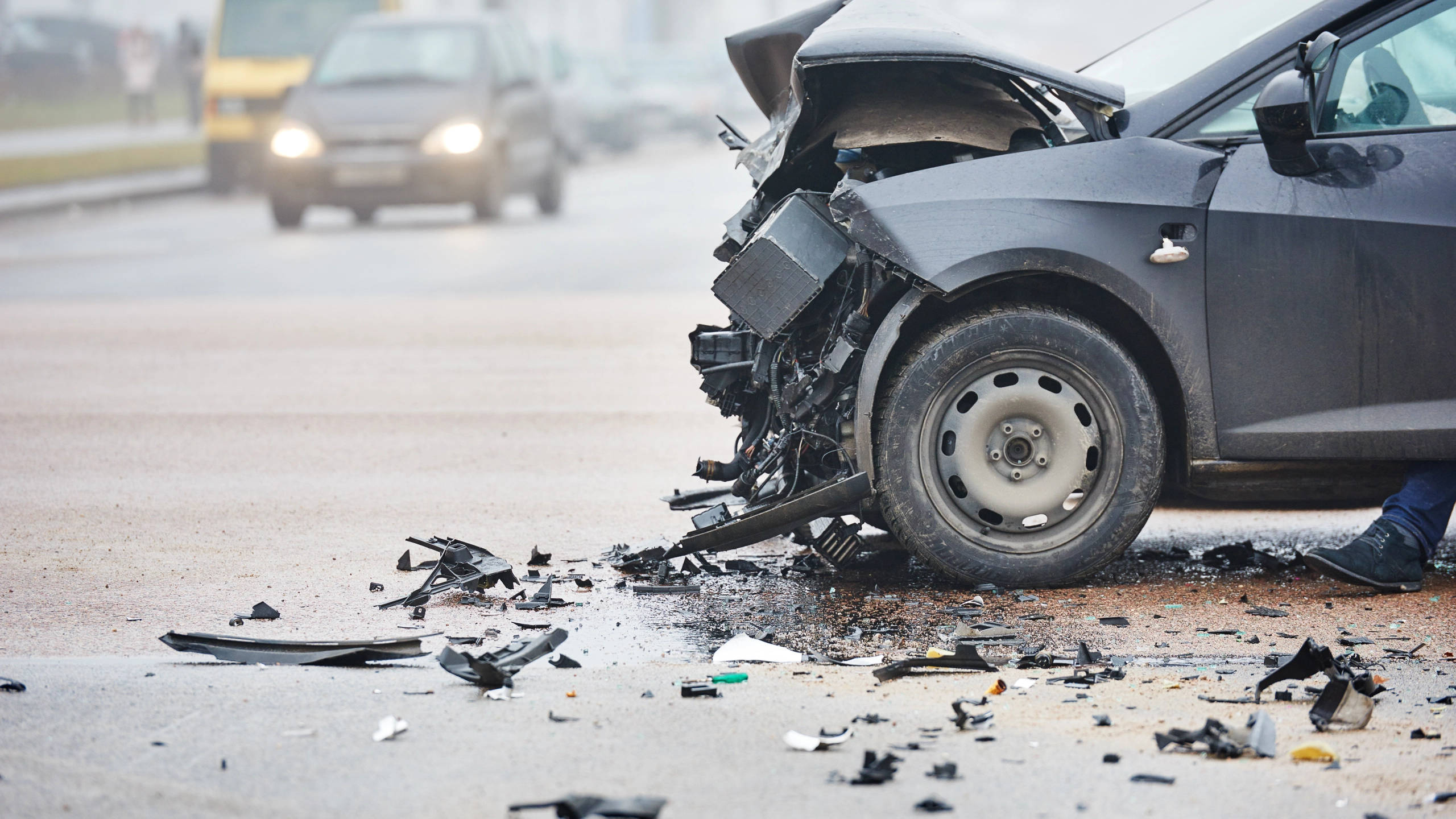 Car Accident 桌布 壁紙 — 一輛嚴重損壞的黑色汽車在潮濕的道路上被撞毀，散落著碎片，輪胎附近可見一個人的腳。