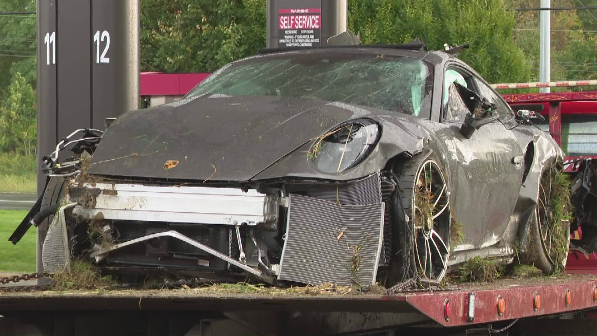 Car Accident 桌布 壁紙 — 一輛嚴重損毀、覆蓋著草叢的黑色保時捷跑車，停放在油站的紅色拖車上。