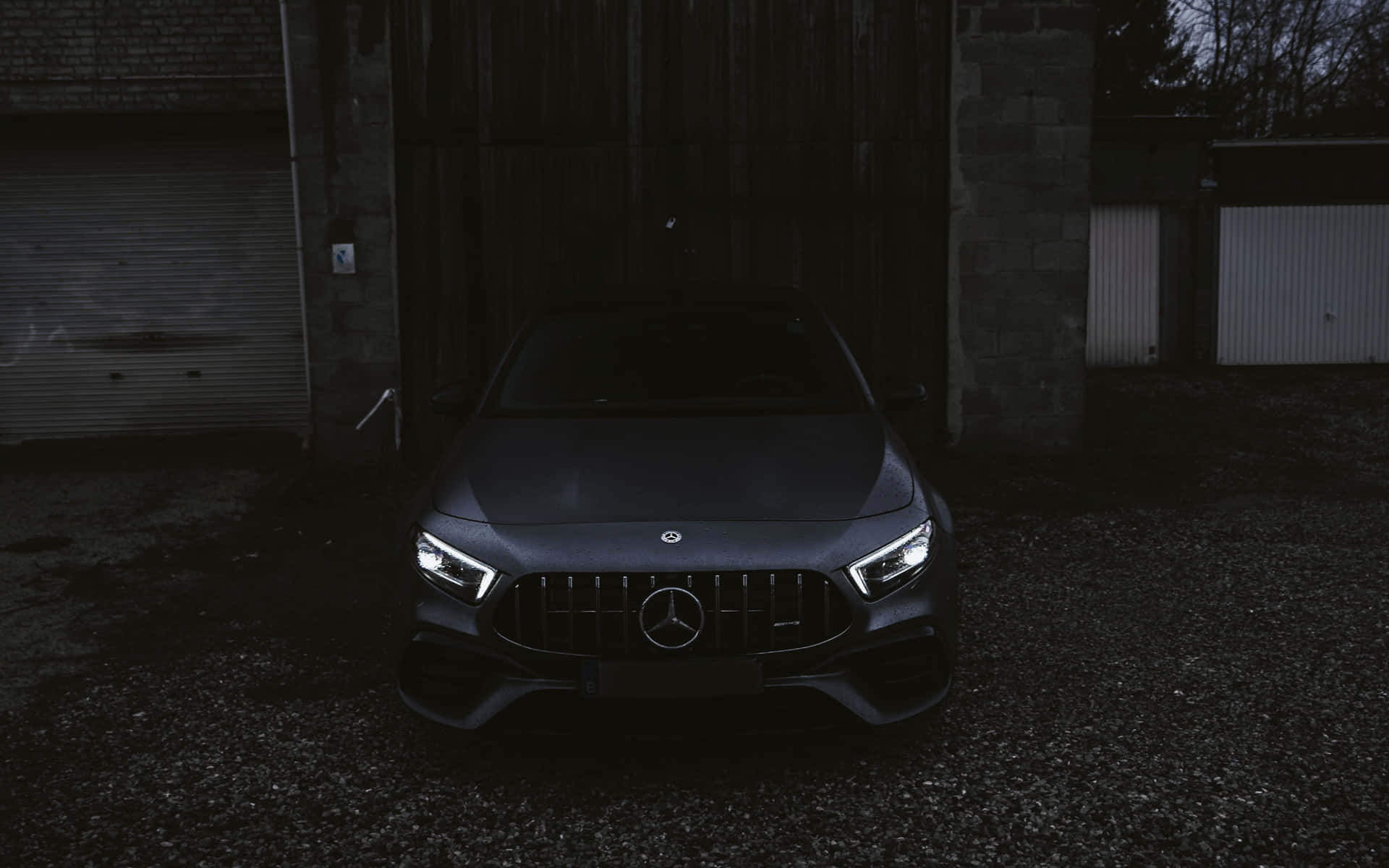 一架深灰色Mercedes-AMG在夜間停泊在碎石庭院中，車頭燈閃爍發光。
