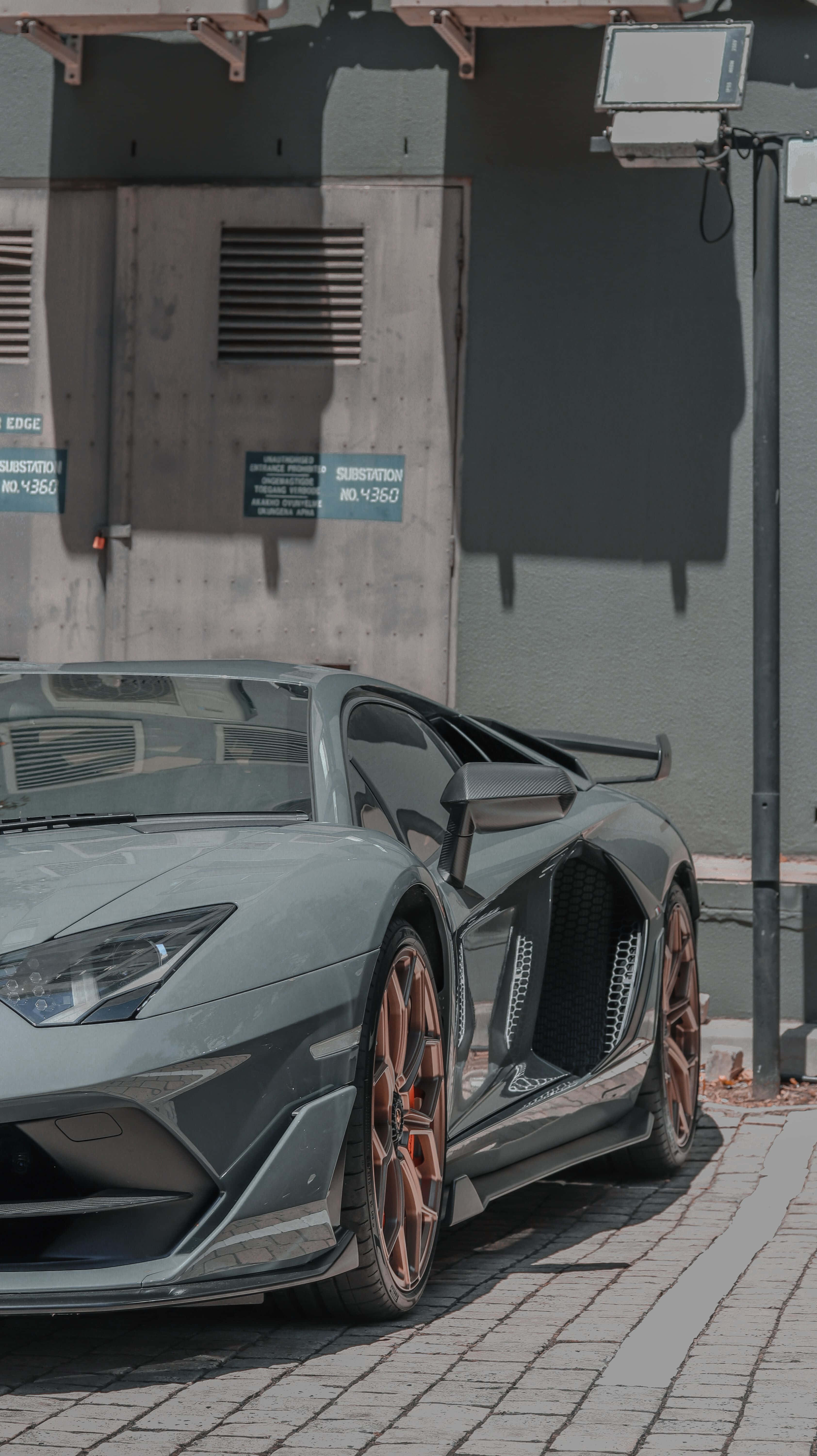 Minimalist Car 桌布 壁紙 — 一輛型格啞光灰色林寶堅尼Aventador配銅色輪圈，停泊在陽光普照的磚砌行人路上，旁邊是混凝土建築物。