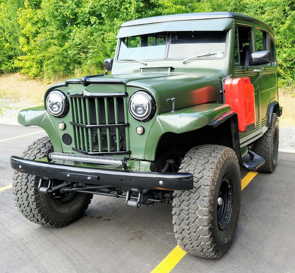 Jeep Willys 桌布 壁紙 — 一輛堅固的橄欖綠色復古吉普車，配備超大輪胎和紅色油桶，停在柏油路上，旁邊是茂密的綠樹。