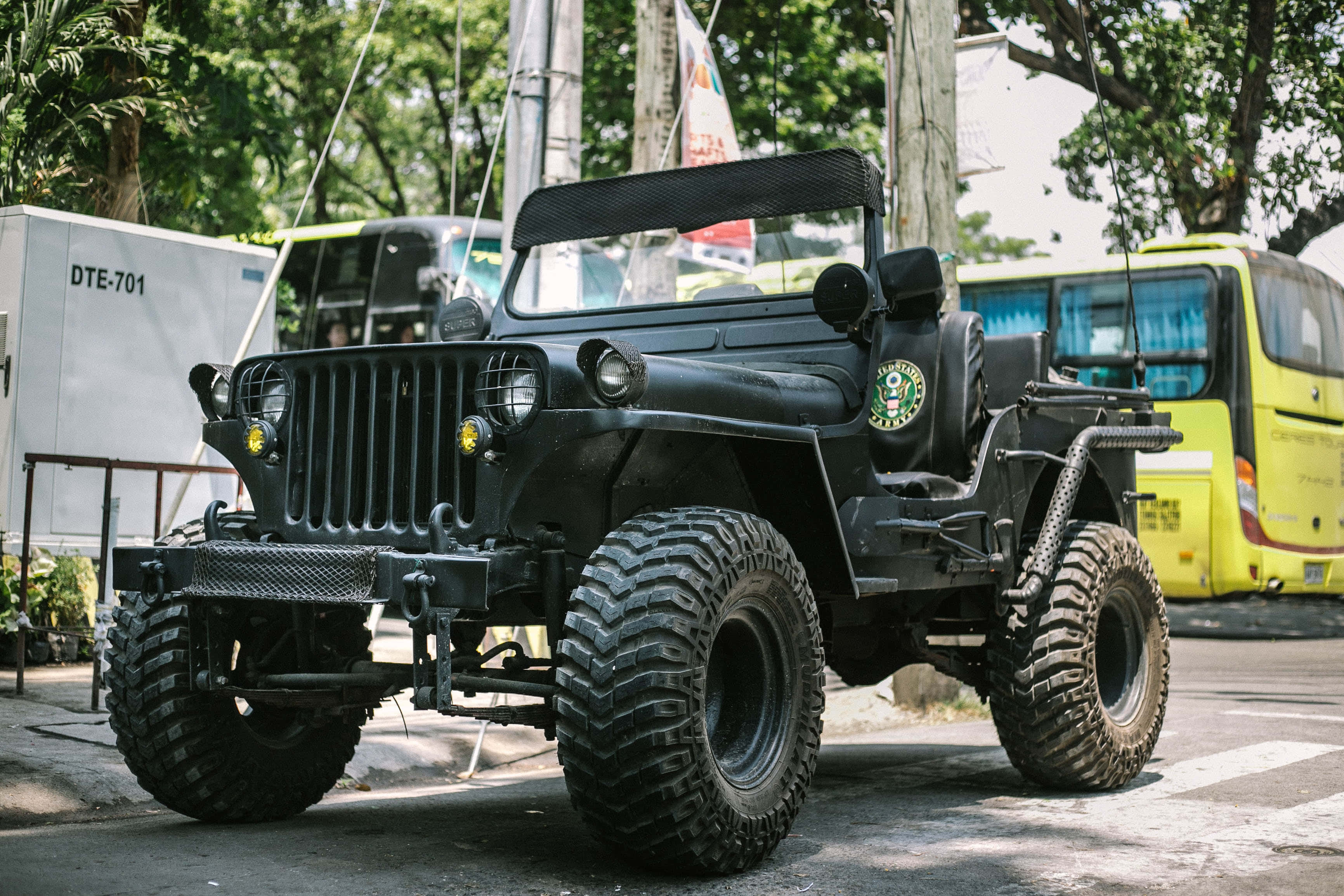 Jeep 動圖 桌布 壁紙 — 一輛堅固的黑色軍用吉普車配備大輪胎，停泊在一條綠樹成蔭的城市街道上，旁邊有一輛黃色巴士。
