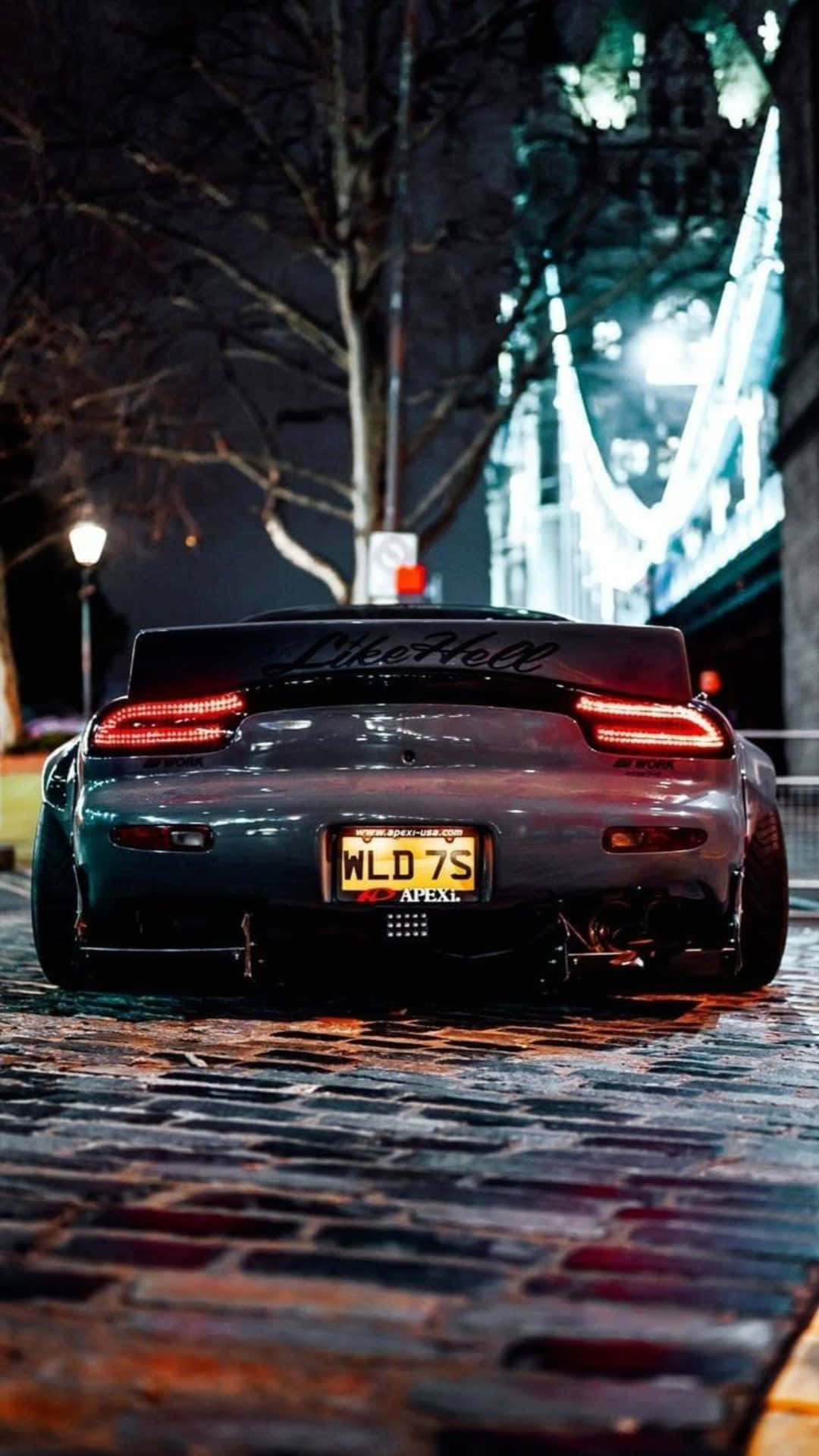 灰色跑車帶紅色尾燈，夜間停在潮濕的鵝卵石街道上，靠近一座亮燈的橋樑。