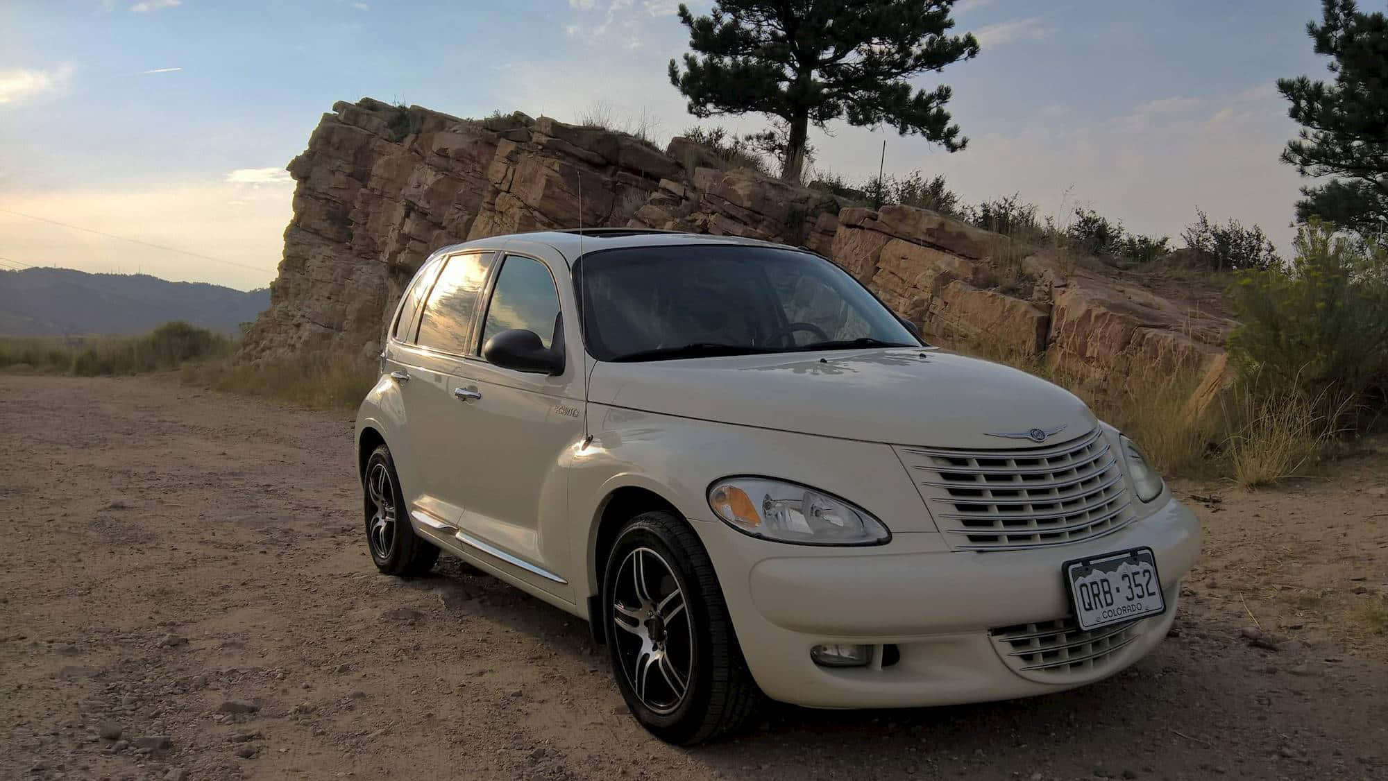 Chrysler Pt Cruiser 桌布 壁紙 — 一輛奶油色的克萊斯勒 PT Cruiser 停泊在一條塵土飛揚的小徑上，旁邊是岩石峭壁，有一棵孤獨的松樹，背景是柔和的日落天空。