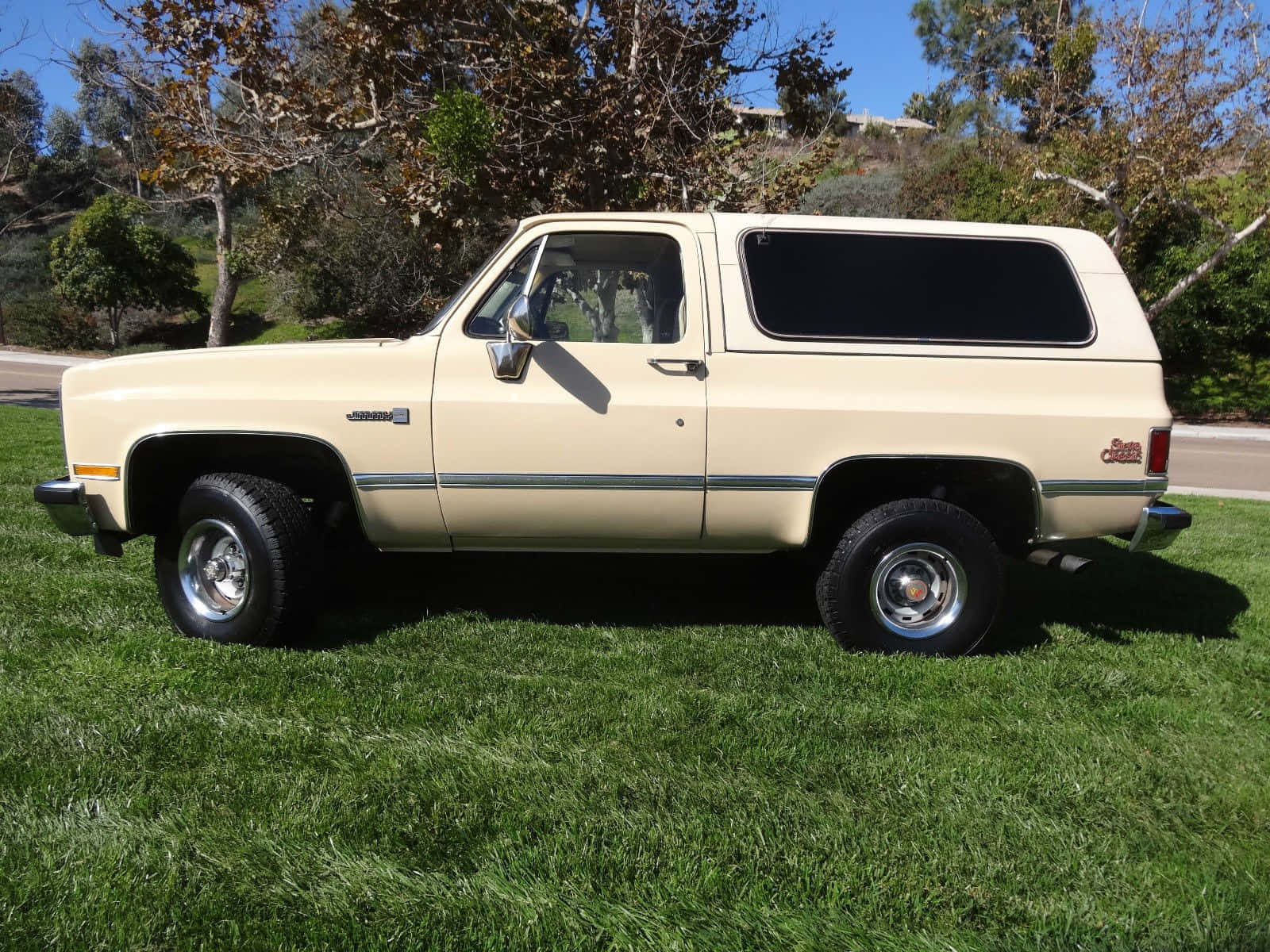Gmc Jimmy 桌布 壁紙 — 一輛奶油色的經典雪佛蘭 Blazer SUV 停放在充滿活力的綠色草地上，背景是清澈的藍天和樹木。