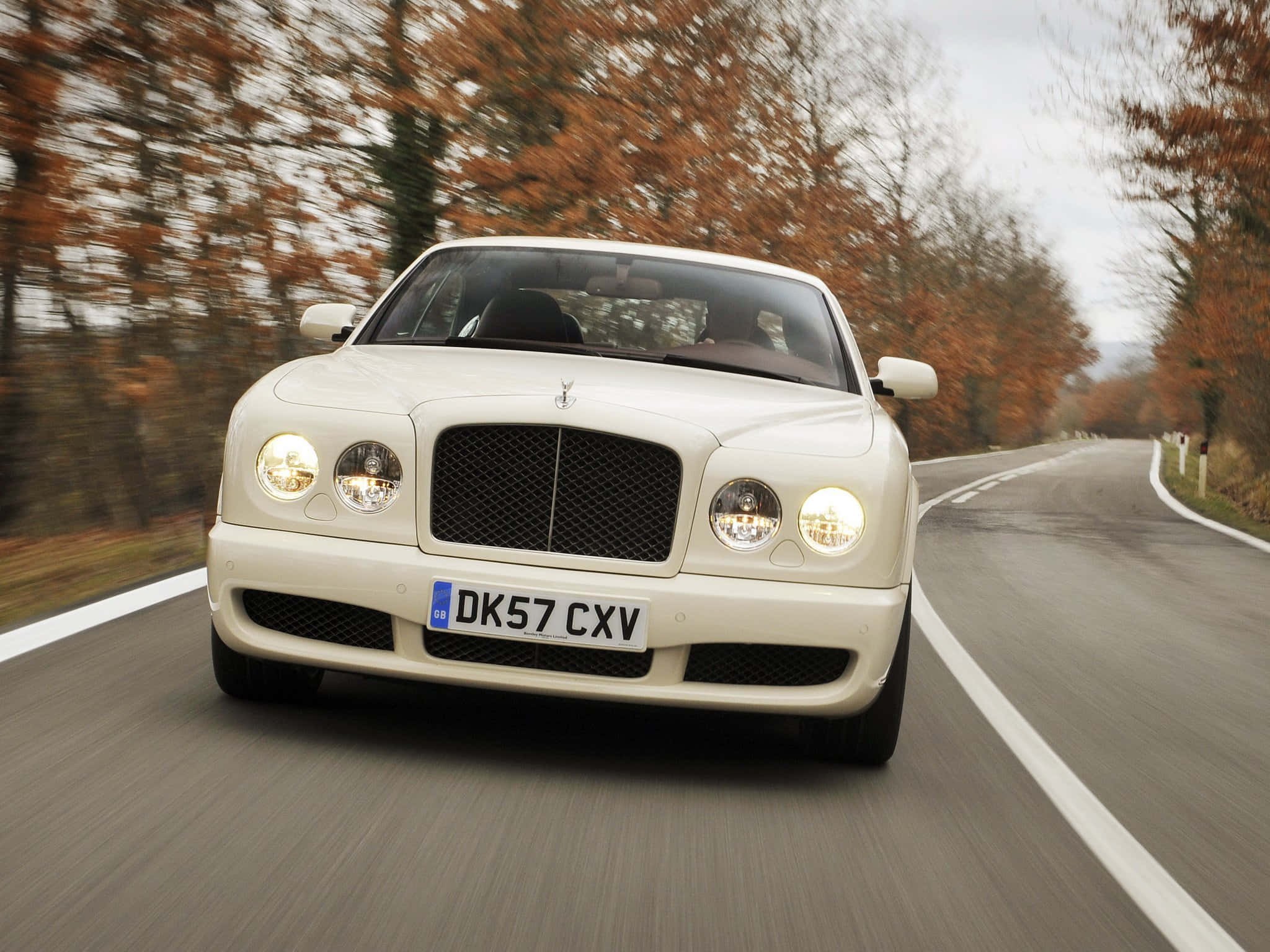 Bentley Brooklands 桌布 壁紙 — 一輛奶油色的賓利 Continental Flying Spur 在陰天之下，沿著兩旁種滿秋樹的蜿蜒道路飛馳。