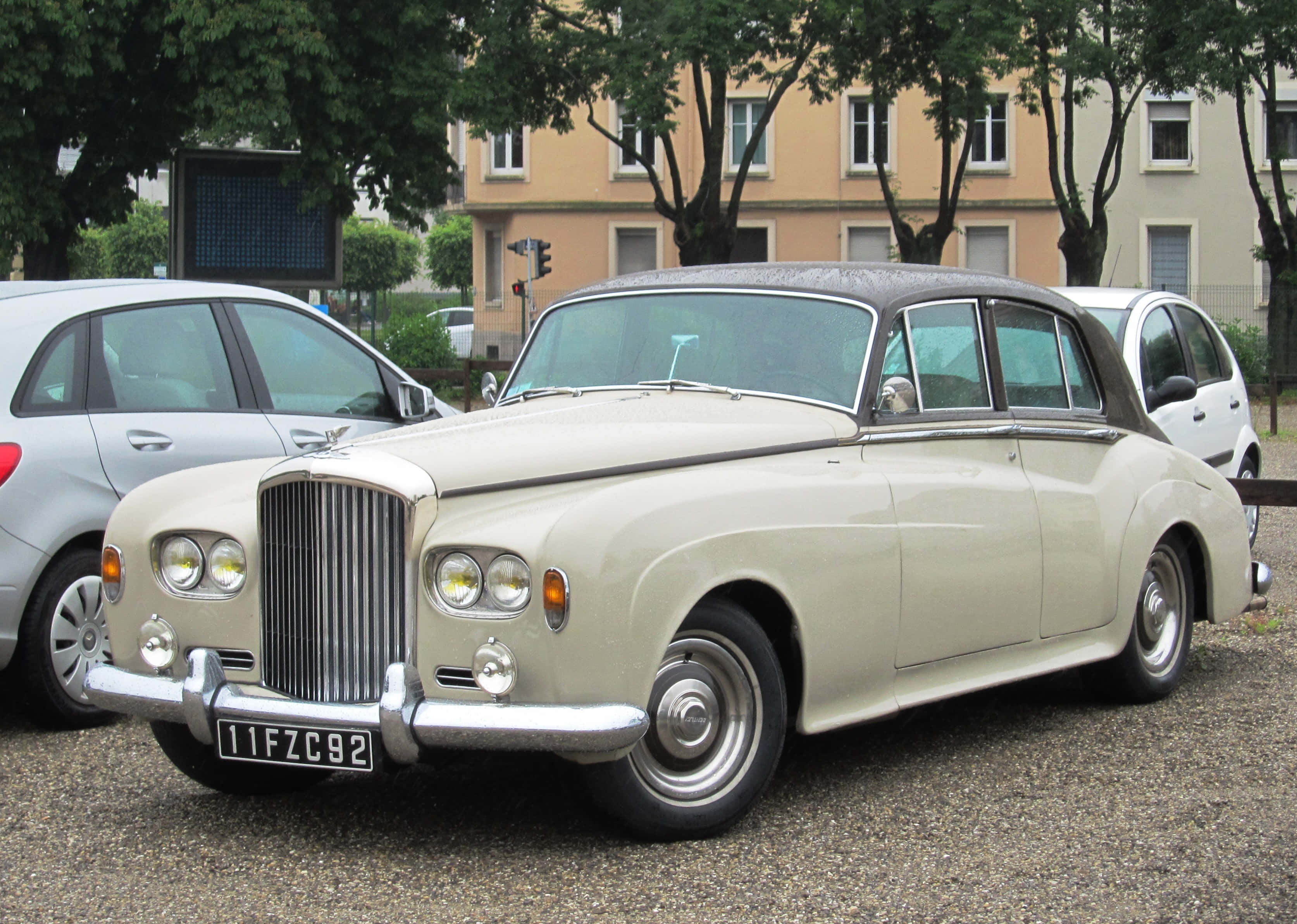 Bentley S3 桌布 壁紙 — 一輛奶油色賓利R-Type轎車，帶有鍍鉻裝飾，停在碎石路上，旁邊是現代汽車，上方有枝葉茂密的樹木。