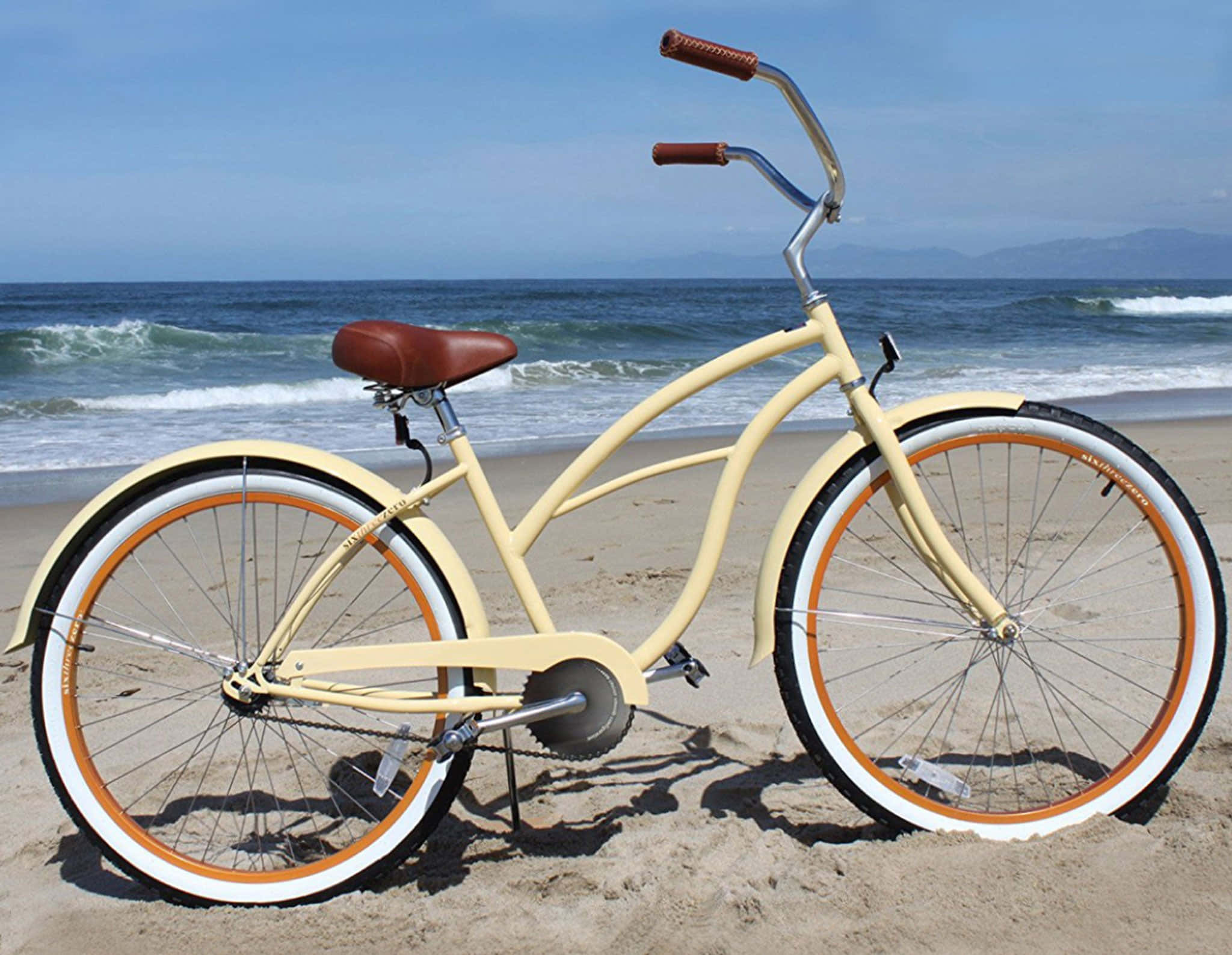 Beach Cruiser 桌布 壁紙 — 一輛奶油黃色沙灘巡洋艦自行車，配有棕色皮革座椅和把手，站在沙灘上，旁邊是清澈藍天下拍打的海浪。