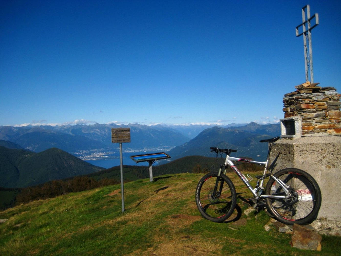 bike riding 桌布 壁紙 — 一輛山地單車靠喺一個草地山頂上嘅石十字架，俯瞰山谷同遠處嘅雪峰，天空晴朗蔚藍。