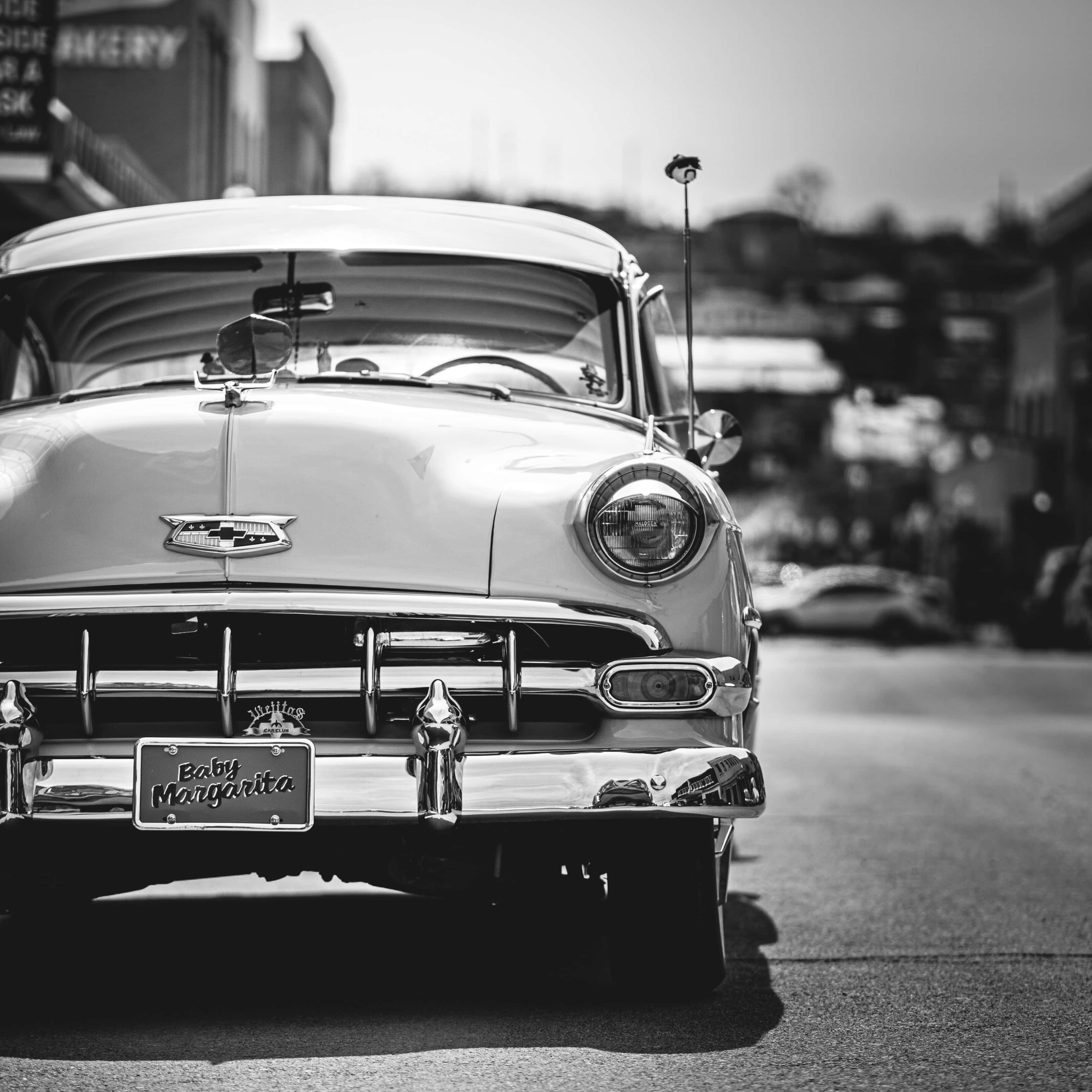 Black And White Vintage Car 桌布 壁紙 — 一輛帶有「Baby Margarita」車牌的復古雪佛蘭汽車在陽光明媚的街道上閃耀著黑白光芒。