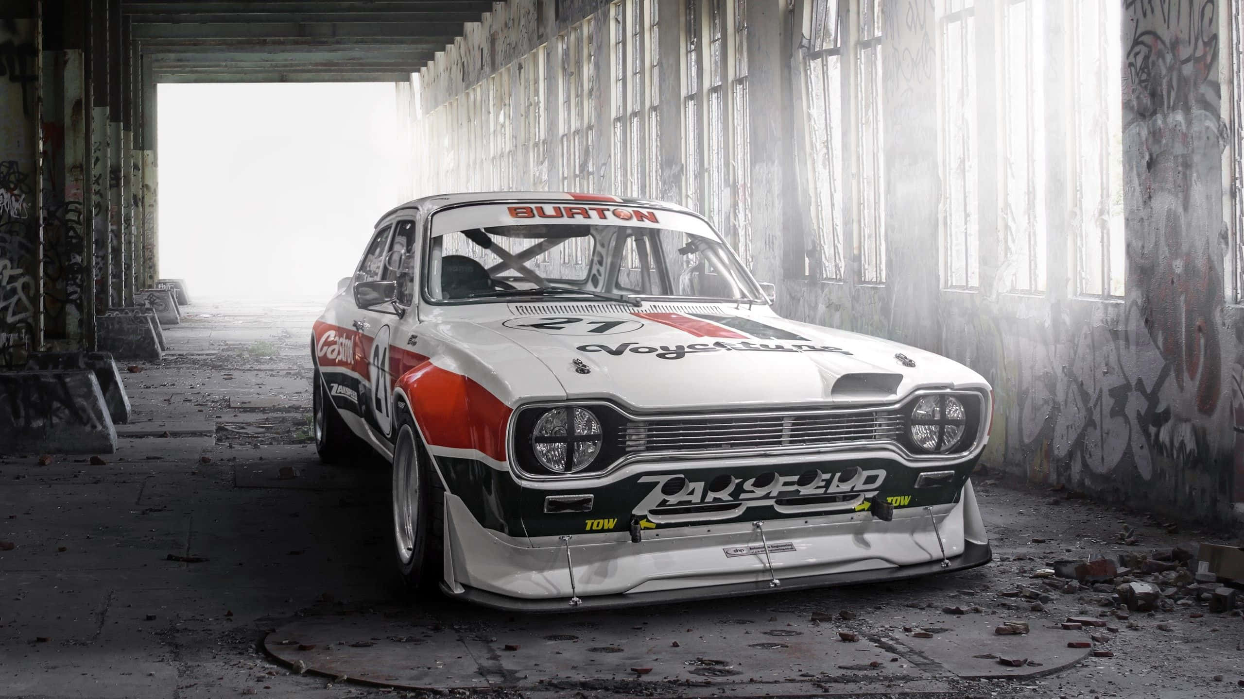 Ford Escort 桌布 壁紙 — 一輛帶有 'Burton' 和 'Oak Speed' 貼紙的復古賽車，戲劇性地停放在一個陽光照射、佈滿塗鴉的廢棄倉庫中。