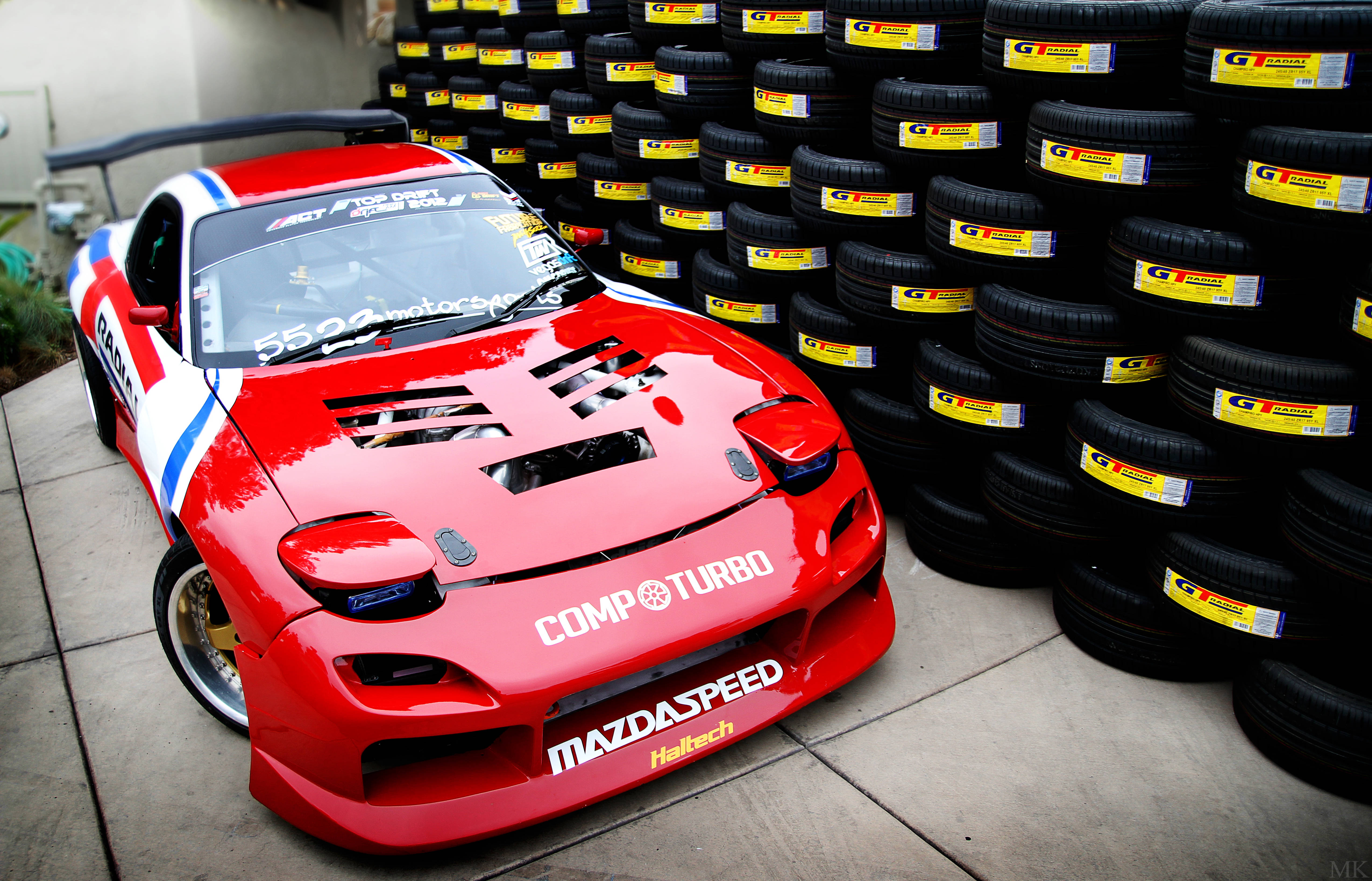 Mazda 桌布 壁紙 — 一輛帶有「CompTurbo」和「MazdaSpeed」貼紙的鮮紅色萬事得RX-7賽車，自豪地站在一堆黑色G Tire賽車輪胎旁邊。