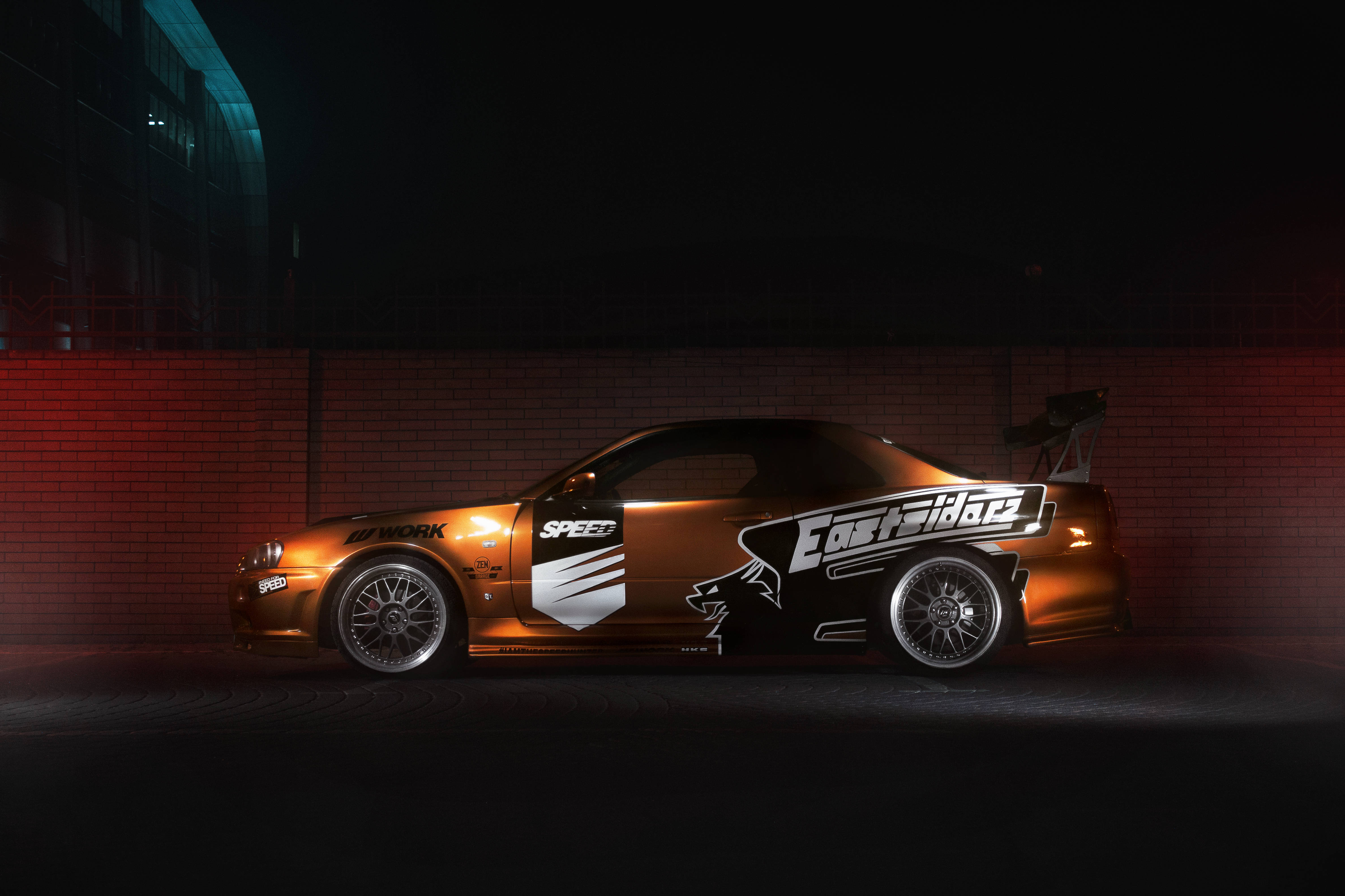 Nissan Skyline GTR R34 Eastsiders 壁紙 — 一輛帶有 'EastRider' 貼紙的金黑色賽車在夜間被戲劇性地照亮，背景是紅磚牆。