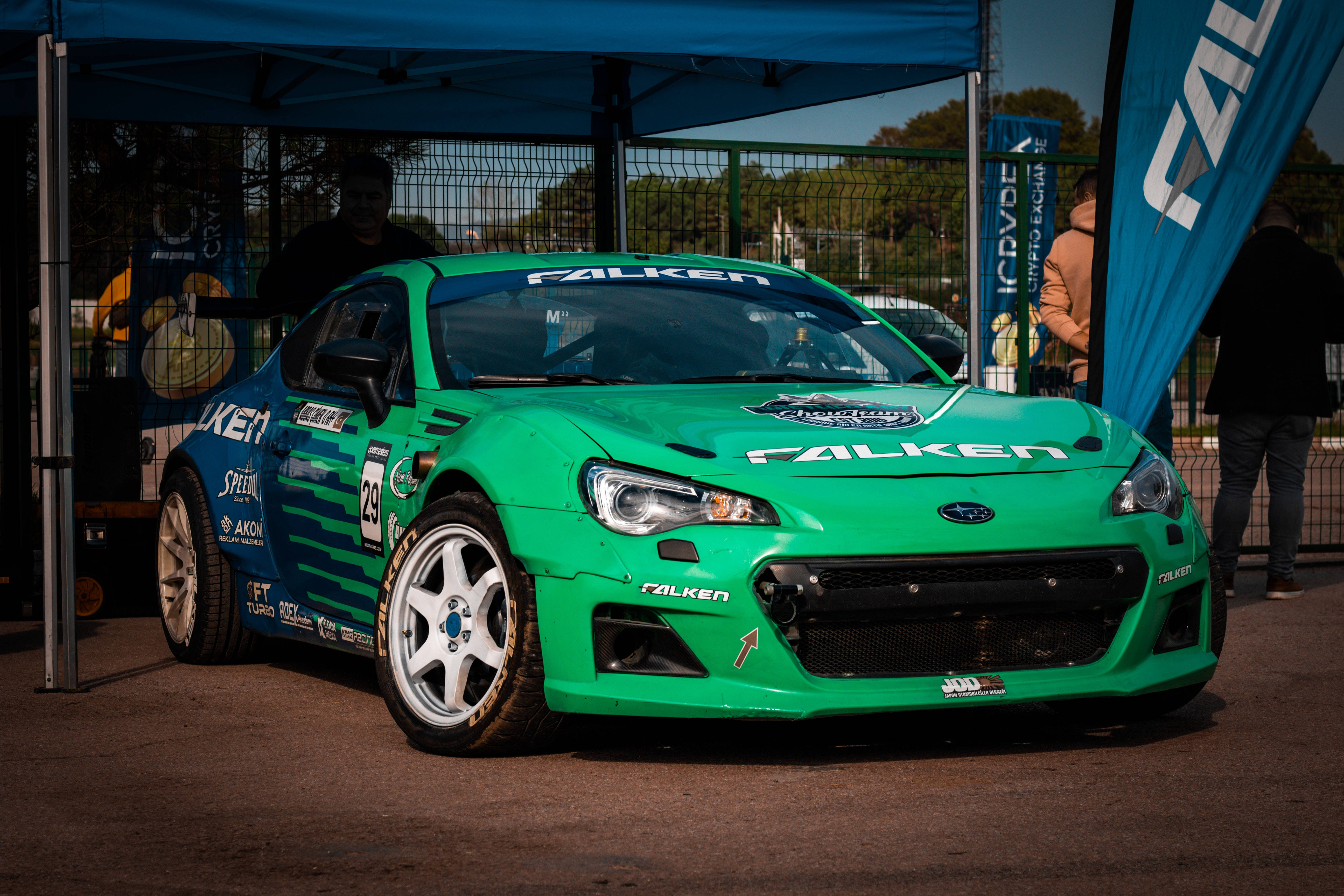 Sports Car 桌布 壁紙 — 一輛帶有 Falken 品牌的鮮綠色賽車停在賽車活動的藍色遮陽篷下，附近有觀眾。